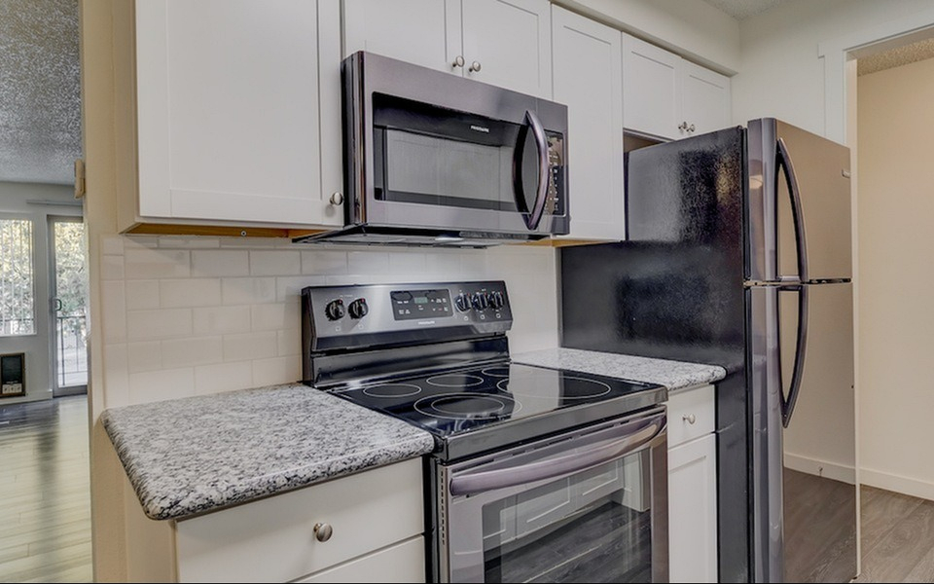 Modern kitchen with stainless steel appliances, granite countertops, and open space leading to a bright living area with large windows.