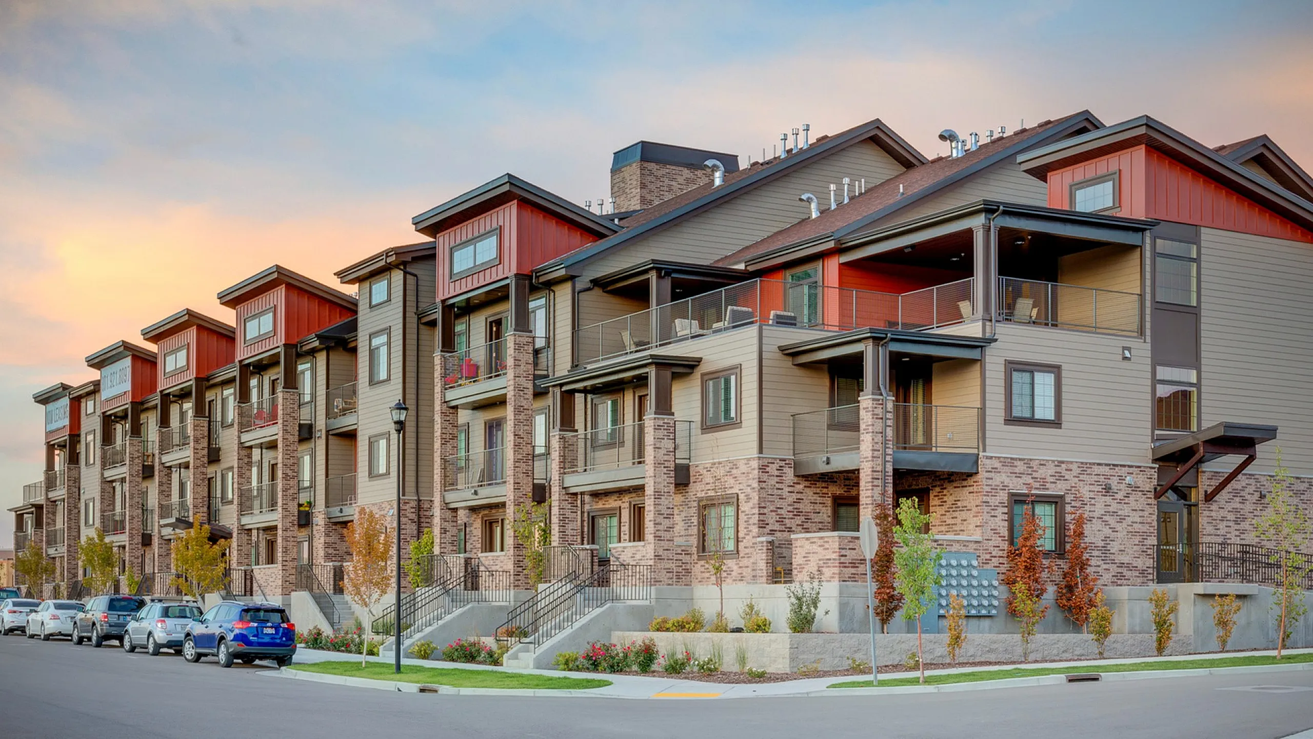 Modern apartment complex with stylish brick and wood facade, featuring spacious balconies and vibrant landscaping at sunset.