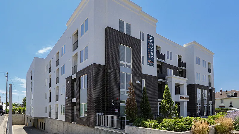 Modern apartment building with sleek black and white exterior, featuring large windows and a welcoming entrance, highlighted by vibrant landscaping under a clear blue sky.