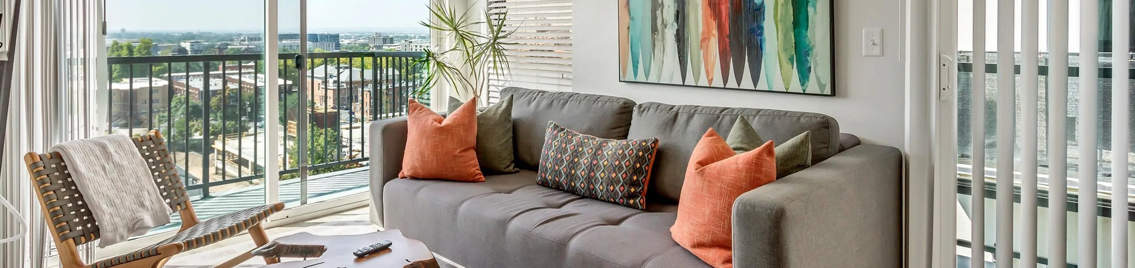 Bright living room with a modern gray sofa adorned with colorful cushions, a unique wooden coffee table, and a chic armchair, all enhanced by floor-to-ceiling windows with cityscape views and stylish abstract wall art.