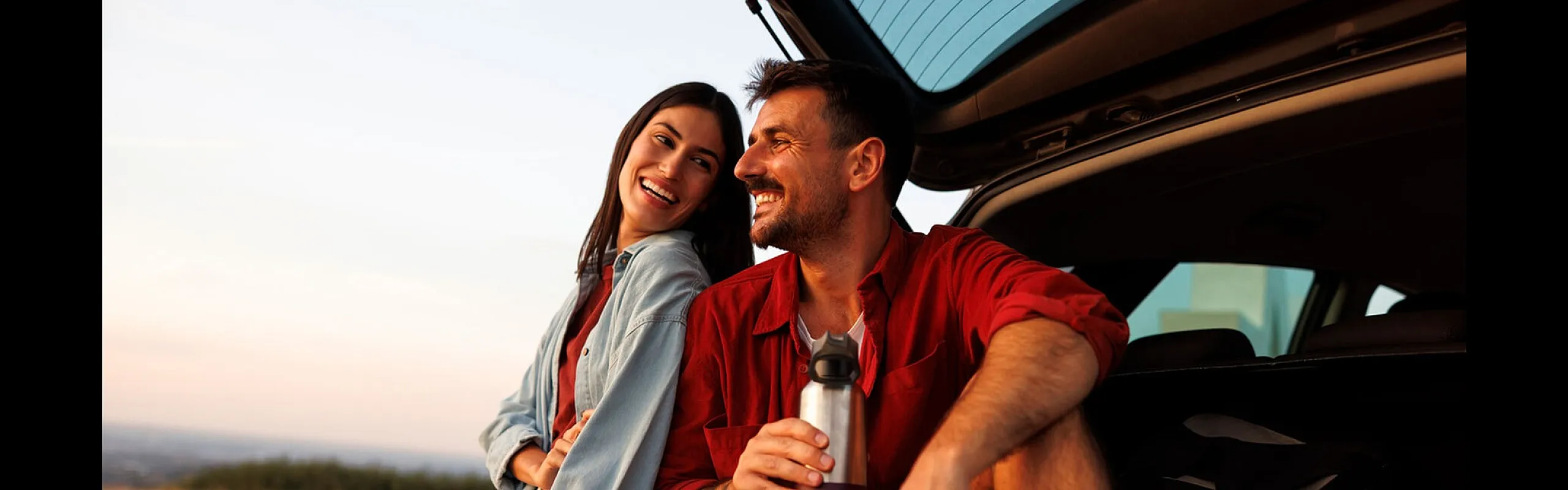 Joyful couple enjoying a scenic sunset on a road trip, sitting by the open trunk of their car in a serene field.