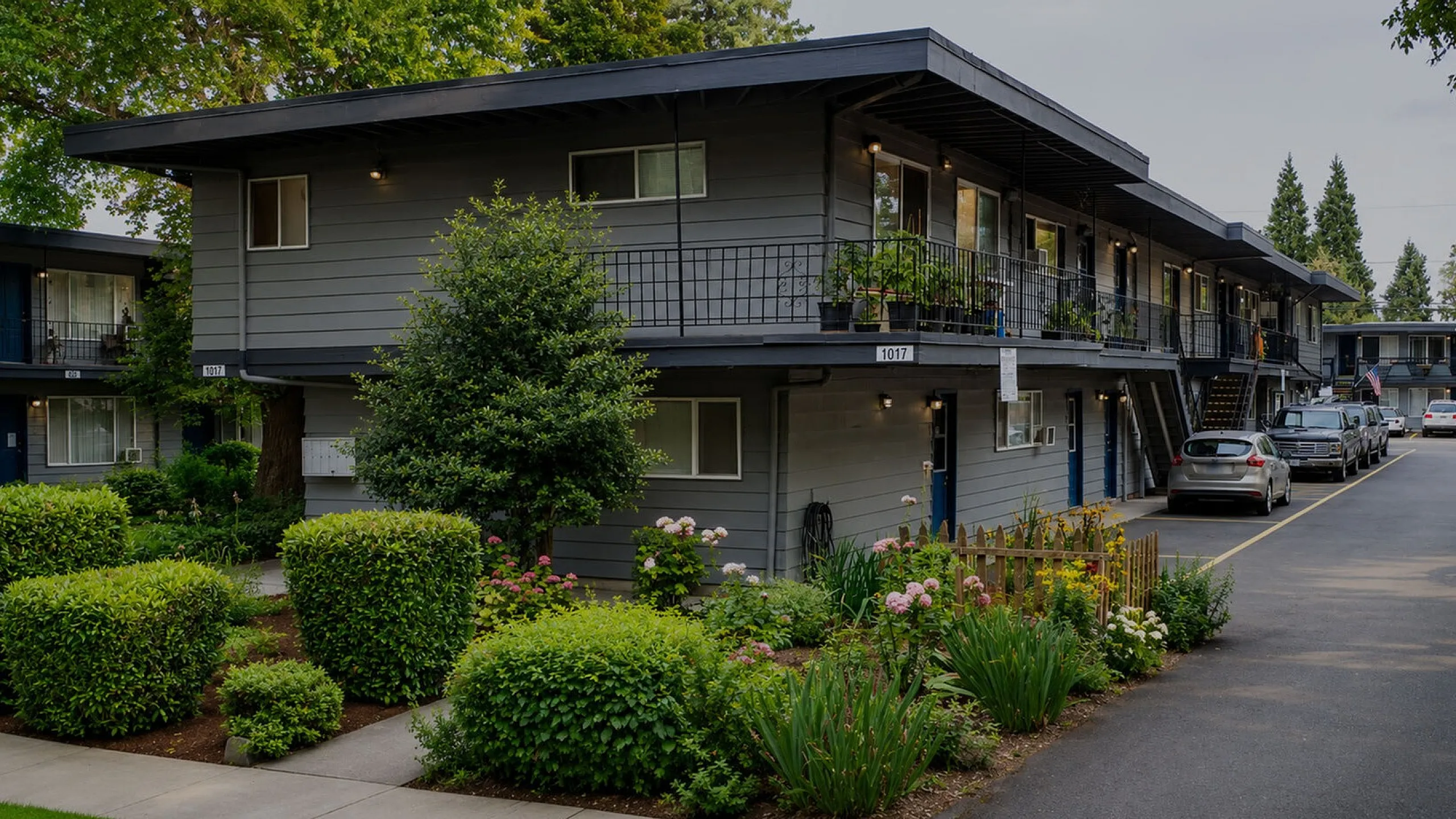 Charming apartment complex with lush landscaping, featuring modern gray exteriors and ample parking space.