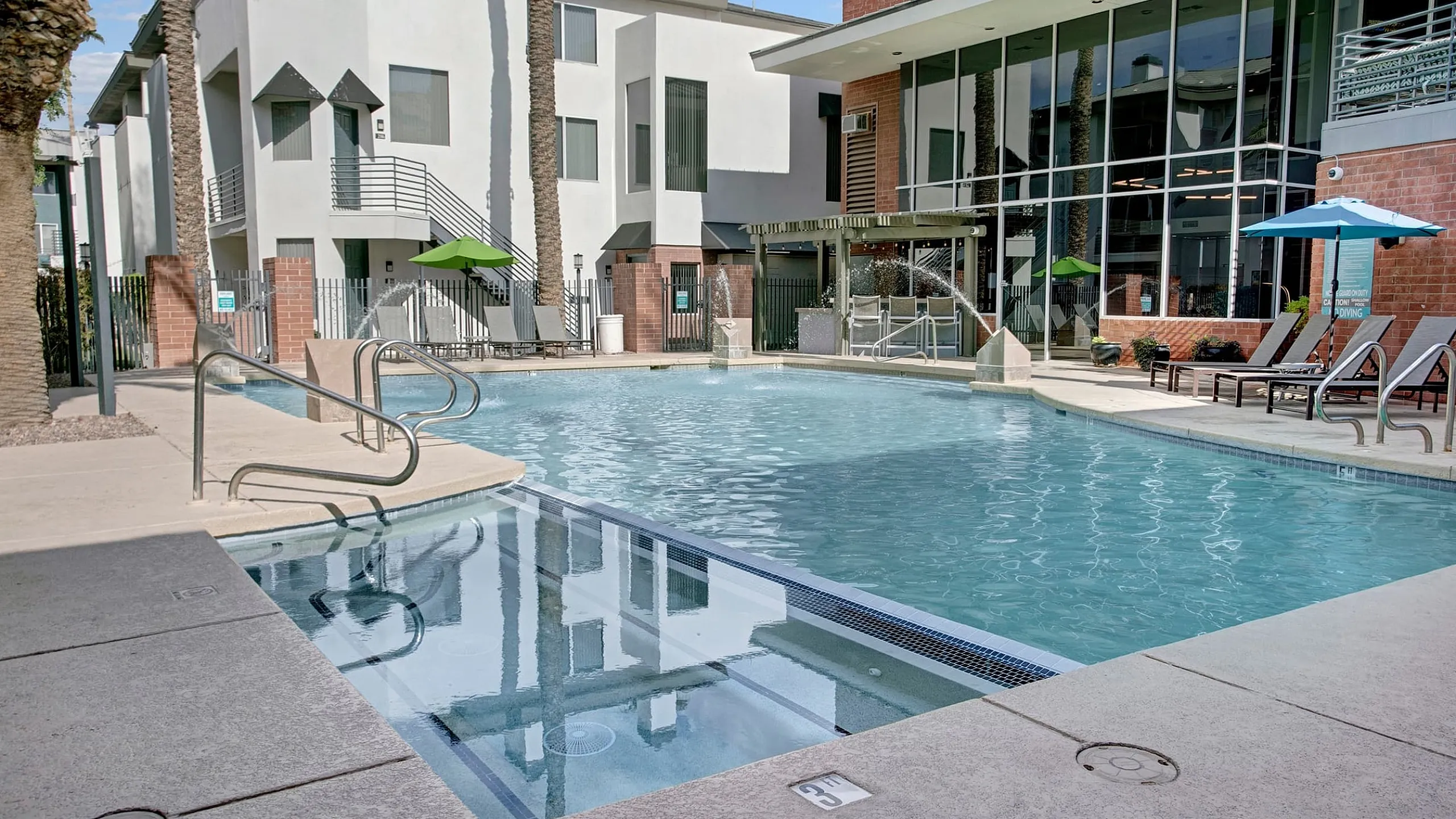 Luxurious apartment complex featuring a sparkling pool with modern architectural design, surrounded by palm trees and inviting lounge chairs.