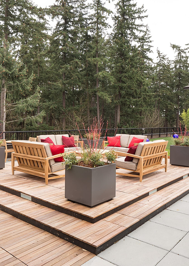 Charming outdoor patio with modern wooden furniture, vibrant red cushions, and lush greenery in a serene natural setting.
