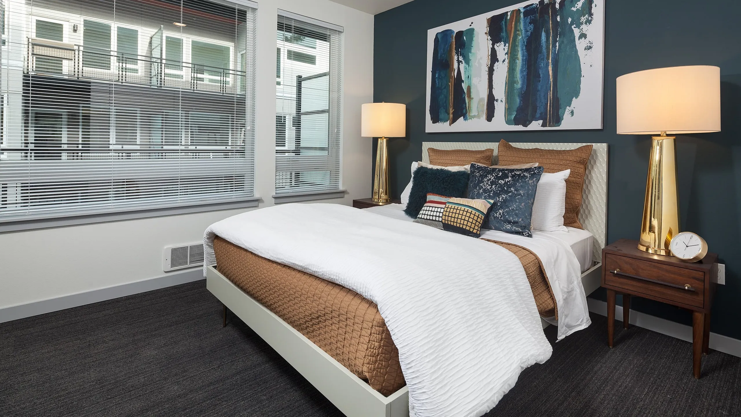Elegant bedroom with modern decor, featuring a cozy bed, stylish lamps, and vibrant artwork on teal accent wall. Large windows offer plenty of natural light, creating a warm and inviting atmosphere.