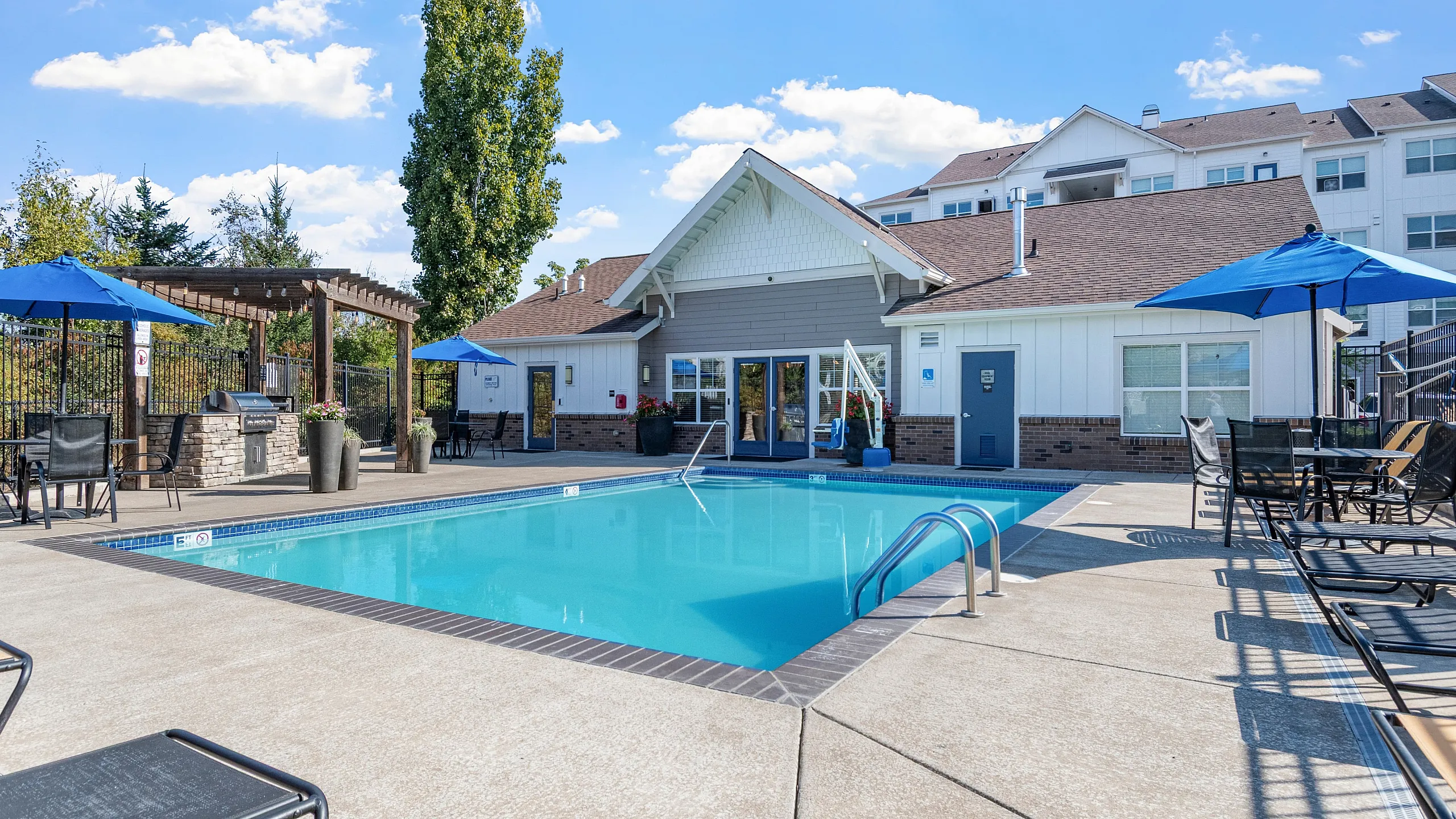 Inviting outdoor pool area with lounge chairs, blue umbrellas, and a charming clubhouse under a vibrant blue sky.