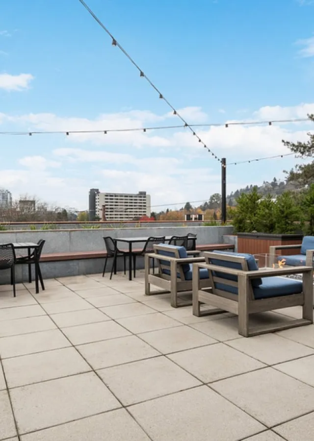 Chic rooftop terrace with comfortable seating, vibrant red flowers, and stunning city skyline views under a bright blue sky.
