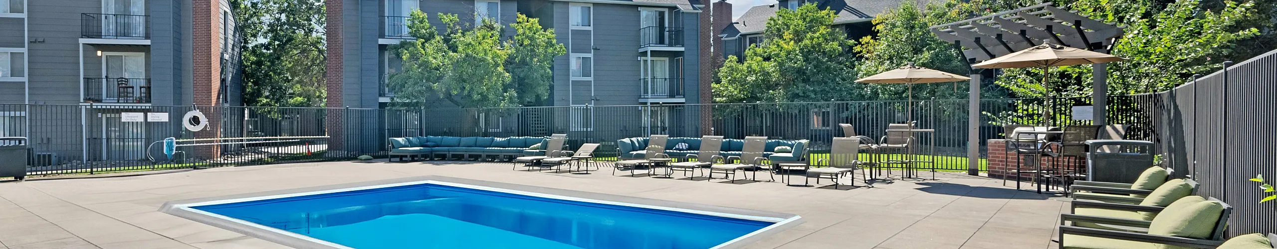 Inviting outdoor pool area with comfortable seating, surrounded by modern apartment buildings and lush landscaping under a clear blue sky.
