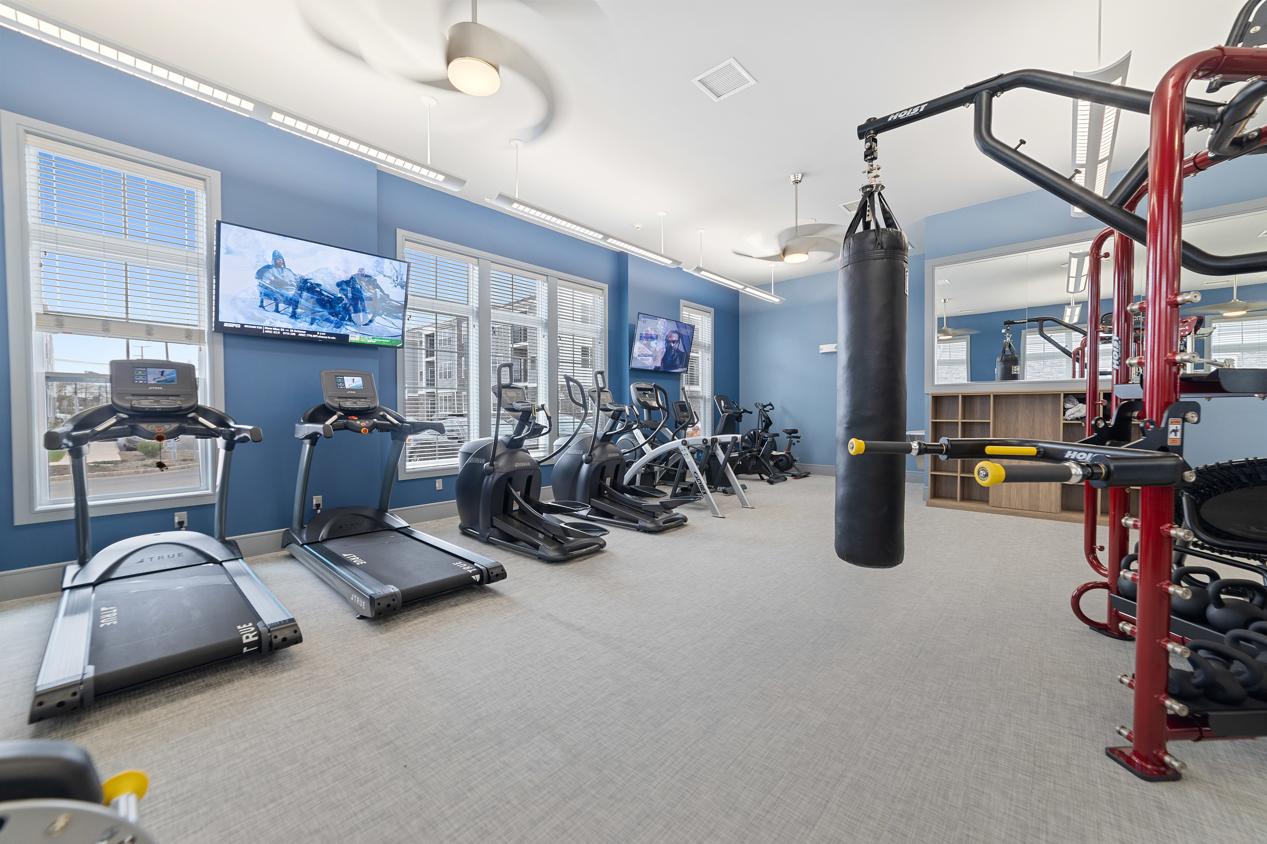 Modern apartment gym with state-of-the-art cardio machines, weightlifting equipment, and flat-screen TVs in a bright, stylish setting.