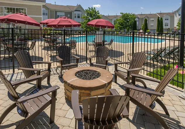 Inviting outdoor space featuring a cozy fire pit surrounded by comfortable chairs, with a view of a sparkling pool and red umbrellas under a clear blue sky.