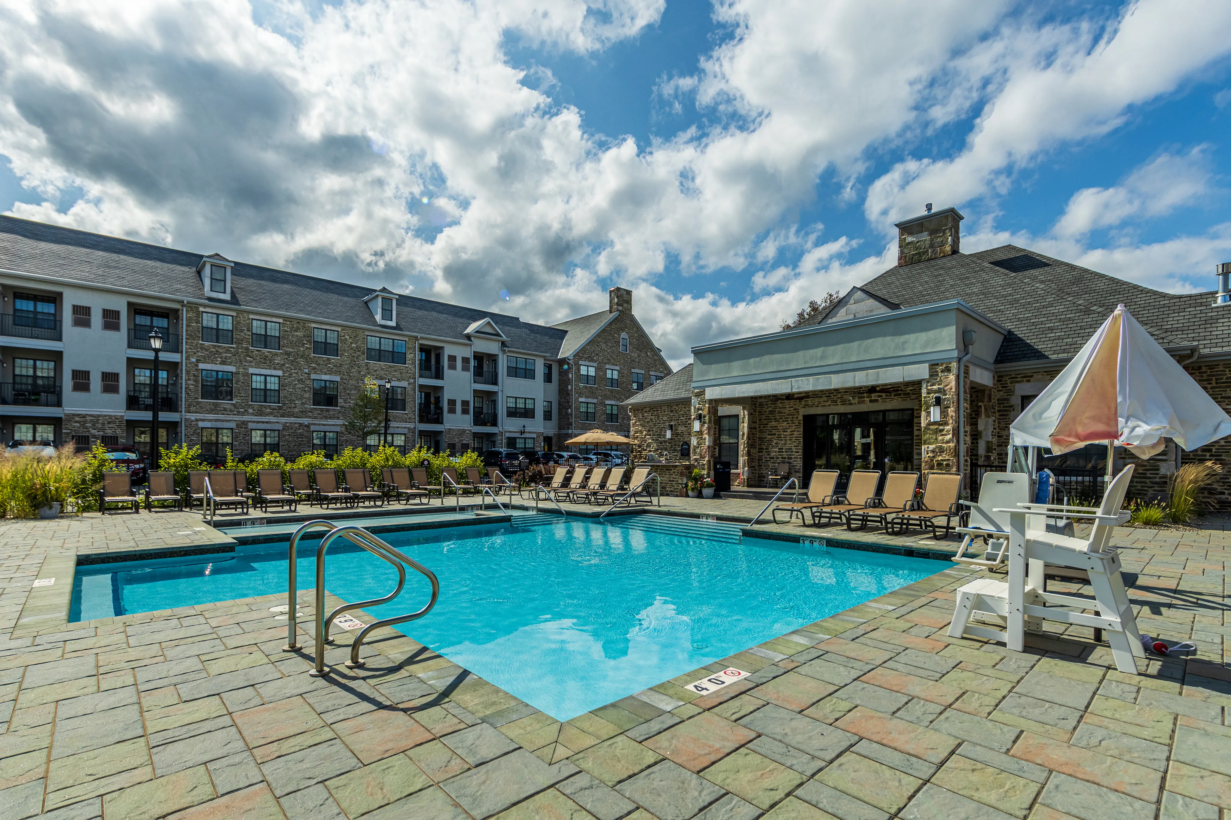 Luxury apartment complex with a stunning outdoor swimming pool, elegant lounge chairs, and charming stone architecture under a vibrant blue sky.