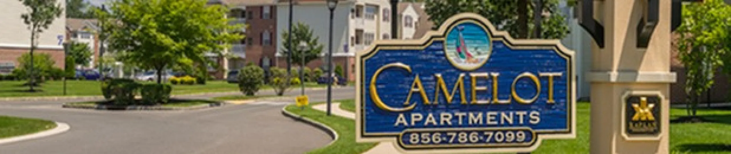 Charming Camelot Apartments entrance with lush greenery, vibrant flowers, and welcoming signage on a sunny day.