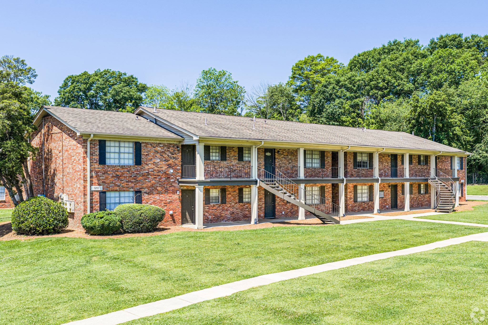Charming brick apartment complex with two stories, surrounded by lush greenery and manicured lawns, offering a comfortable living space with natural light and inviting pathways.