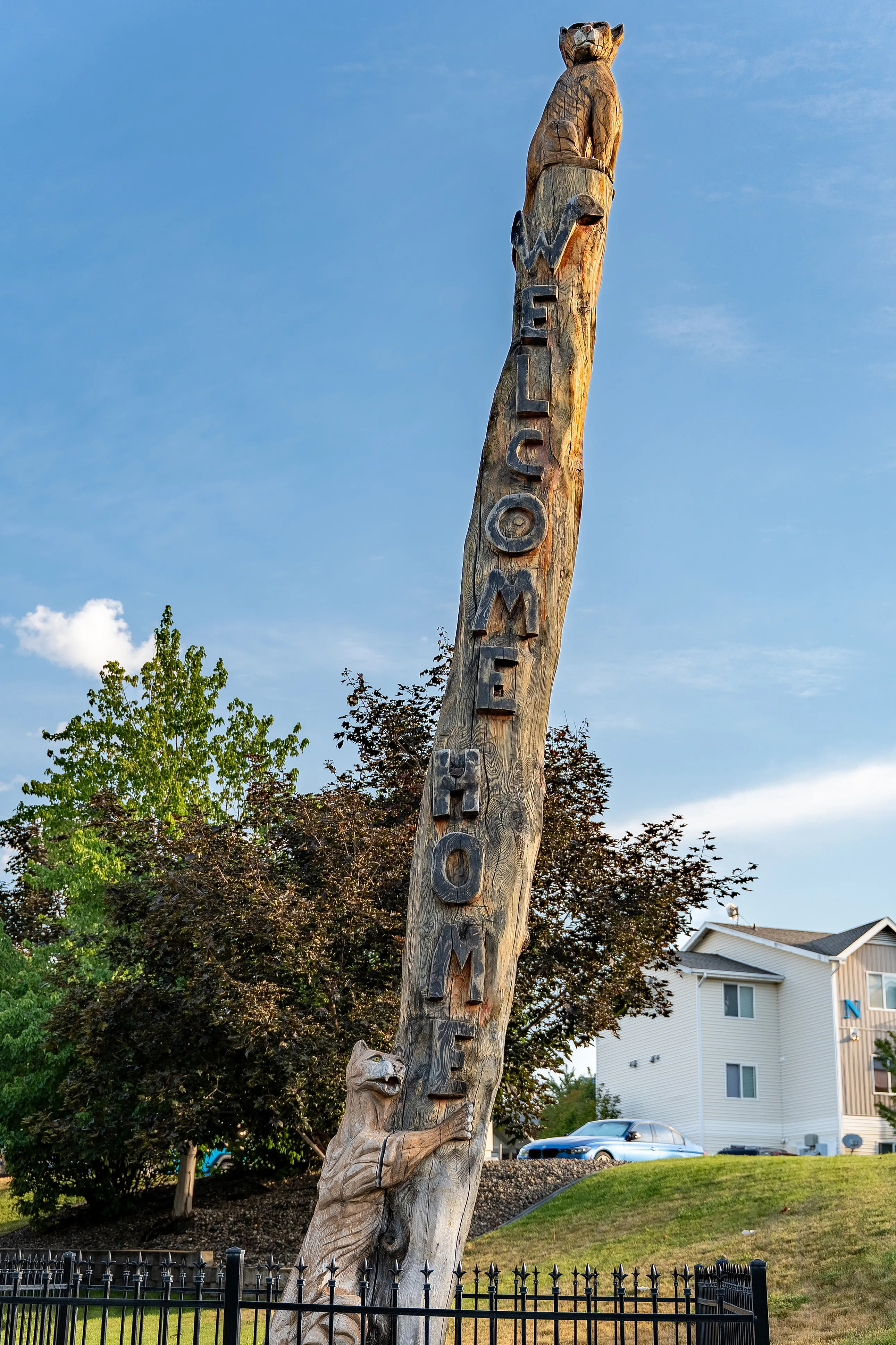 Charming welcome home sign carved into a tall tree trunk with playful animal sculptures, set against a backdrop of clear blue sky and lush greenery.