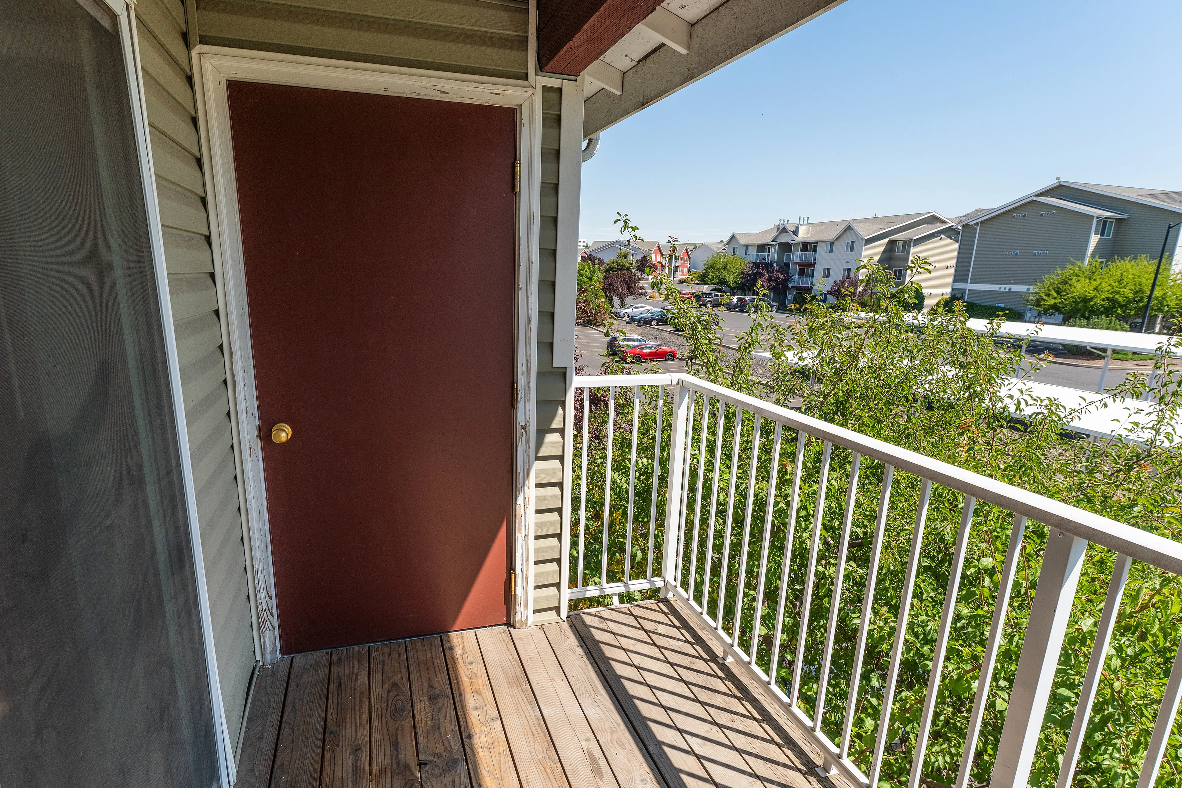 Charming apartment balcony with wooden flooring, red door, and scenic views of a vibrant neighborhood and greenery.