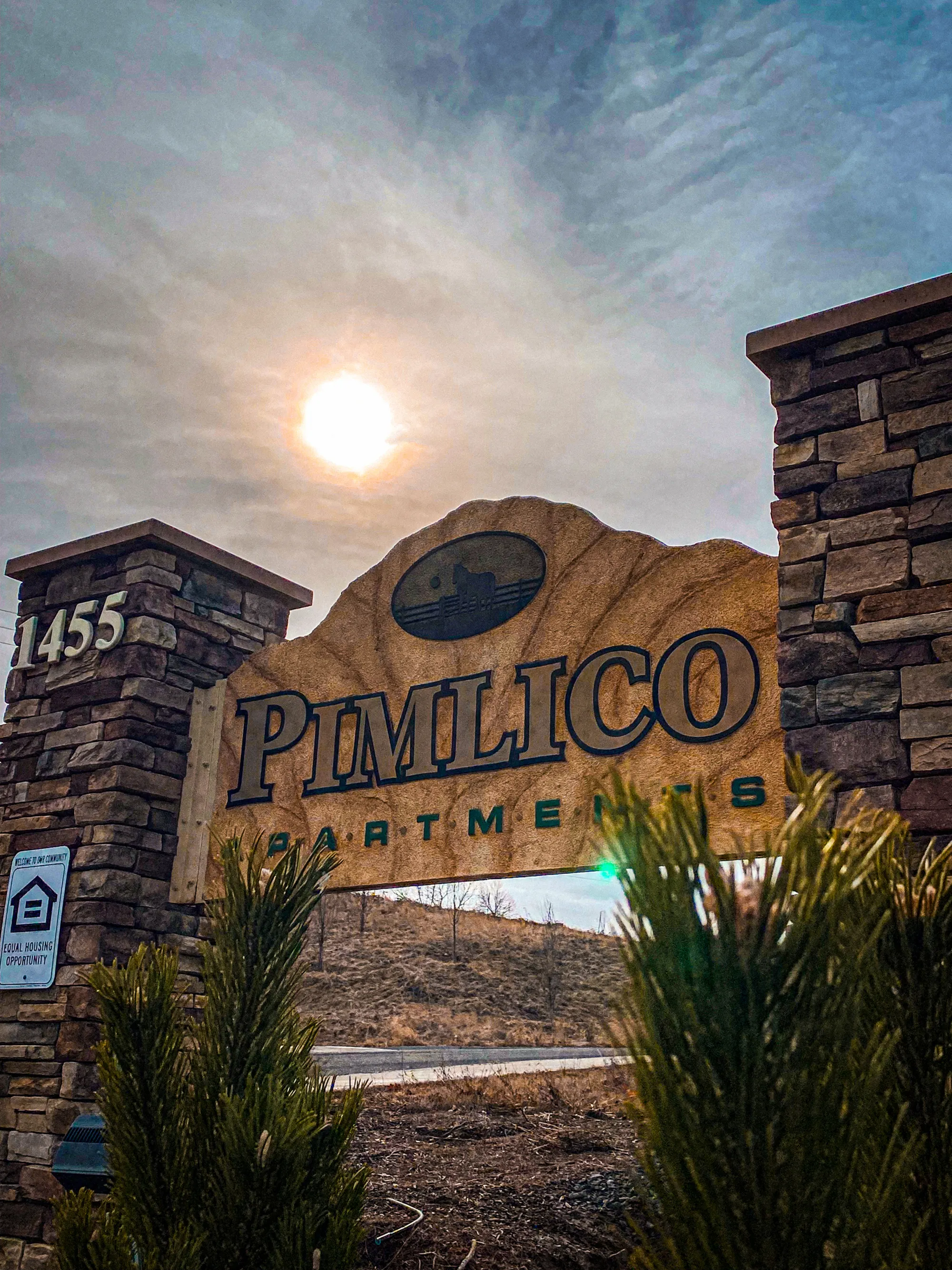 Inviting entrance of Pimlico Apartments featuring a rustic stone sign basking in the warm sunlight, framed by lush greenery.