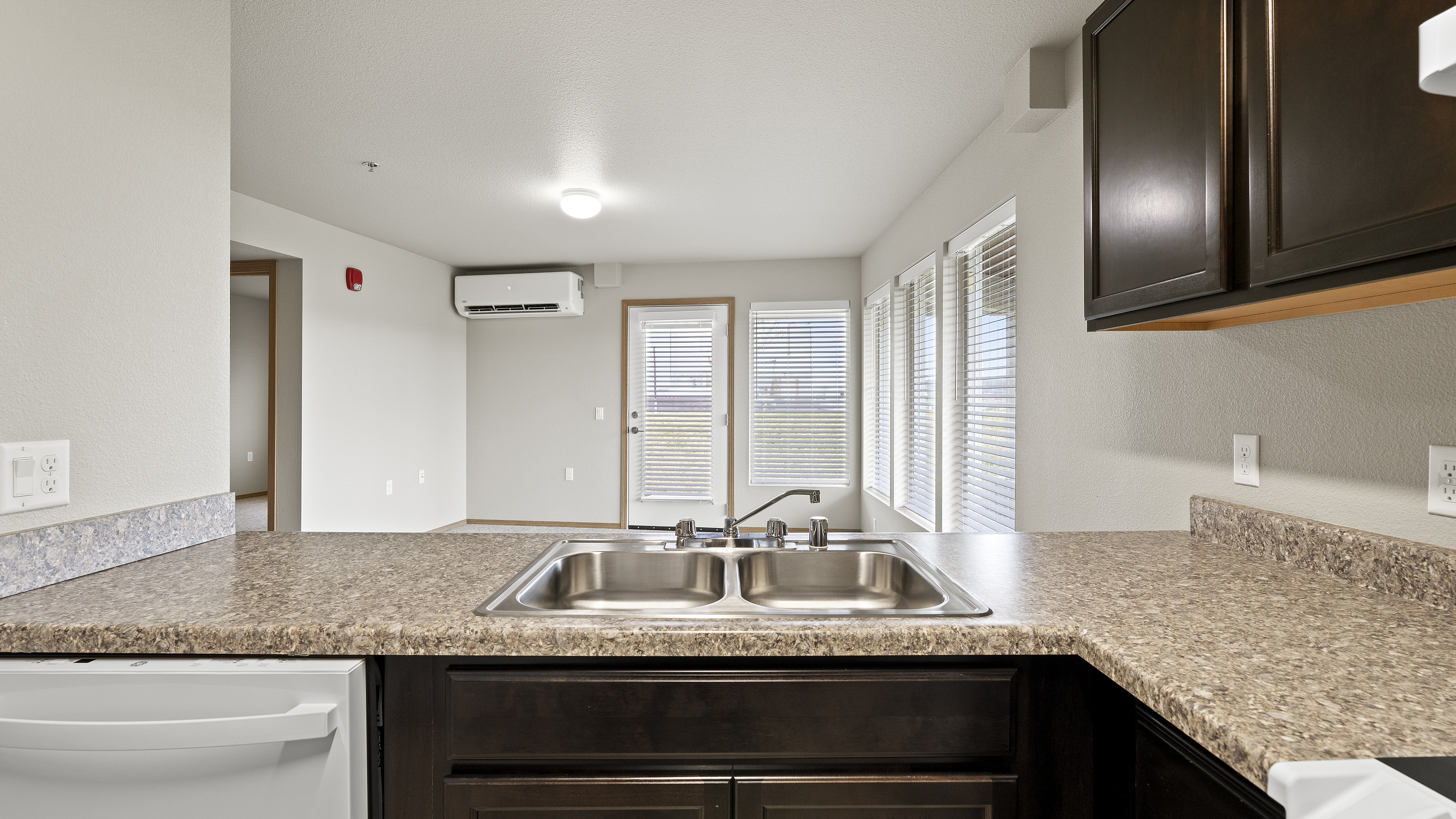 Bright and modern open-plan kitchen with granite countertops and stainless steel sink, leading to a sunlit living area with large windows.