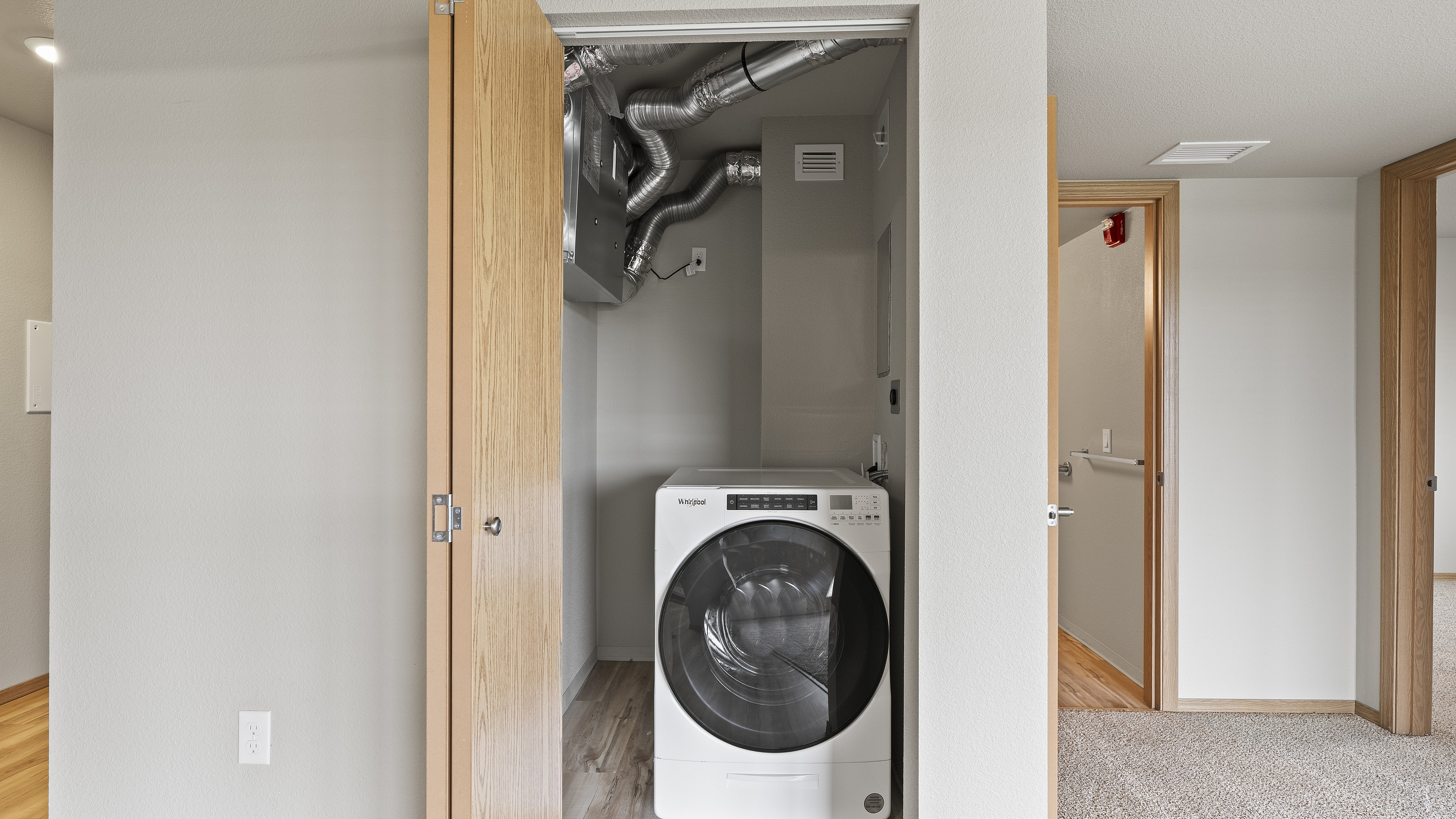 Modern apartment with a convenient laundry nook featuring a sleek, front-loading washer, adjacent to spacious carpeted hallways.