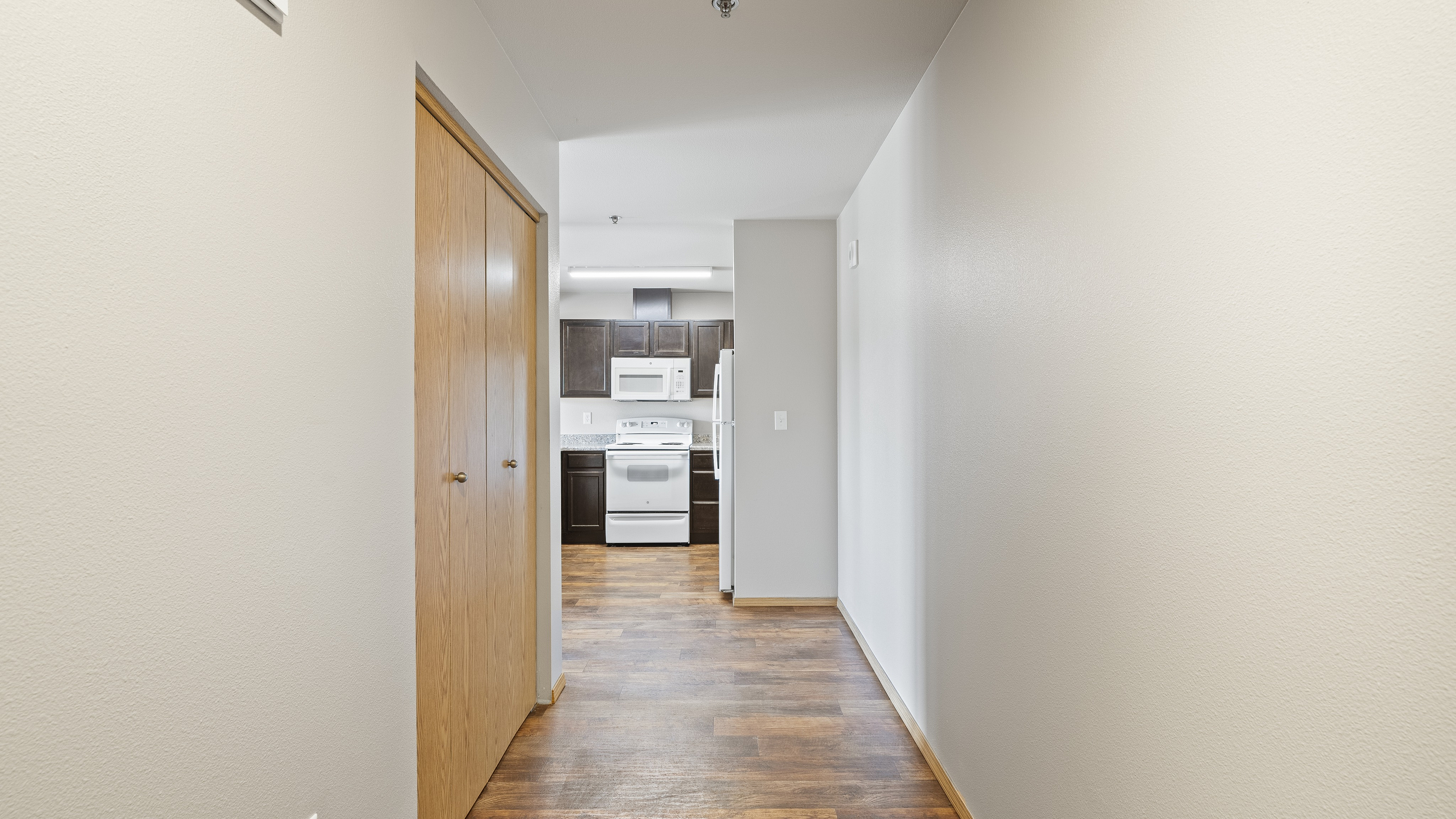 Inviting apartment hallway leading to a modern kitchen with dark cabinets and white appliances, featuring wood-style flooring.