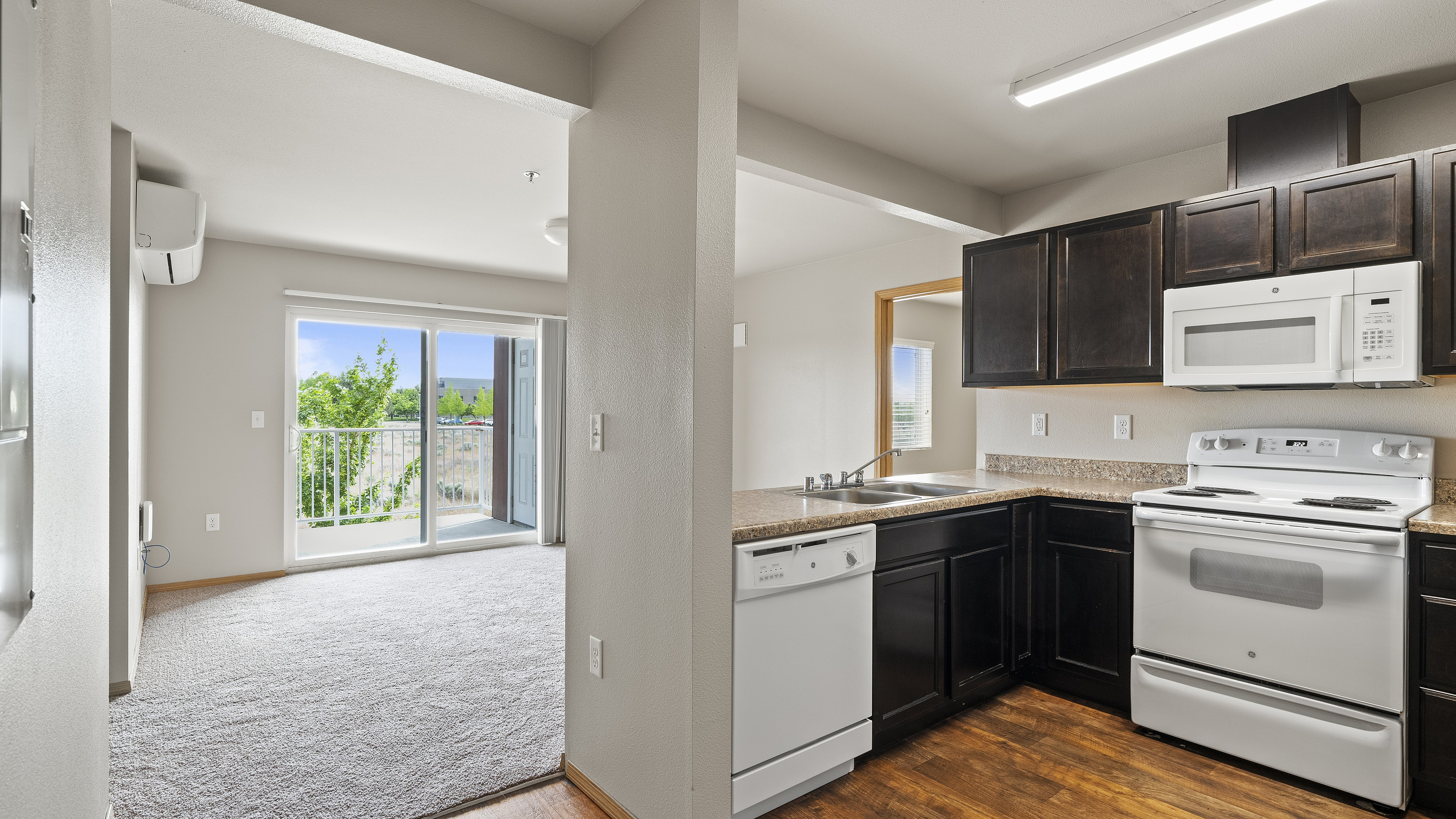 Bright and inviting apartment interior showcasing a modern kitchen with dark cabinets and white appliances, adjacent to a carpeted living room with balcony access and scenic views.