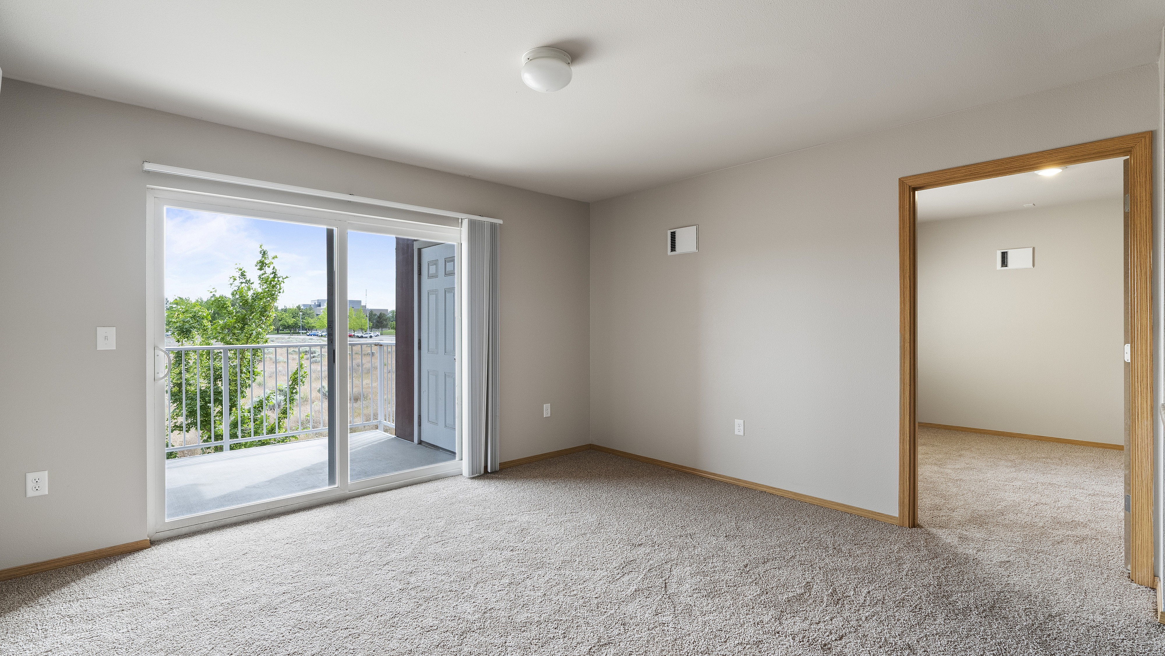 Bright living room with a cozy carpet floor, large sliding glass doors opening to a scenic balcony, and a view into the adjoining room.