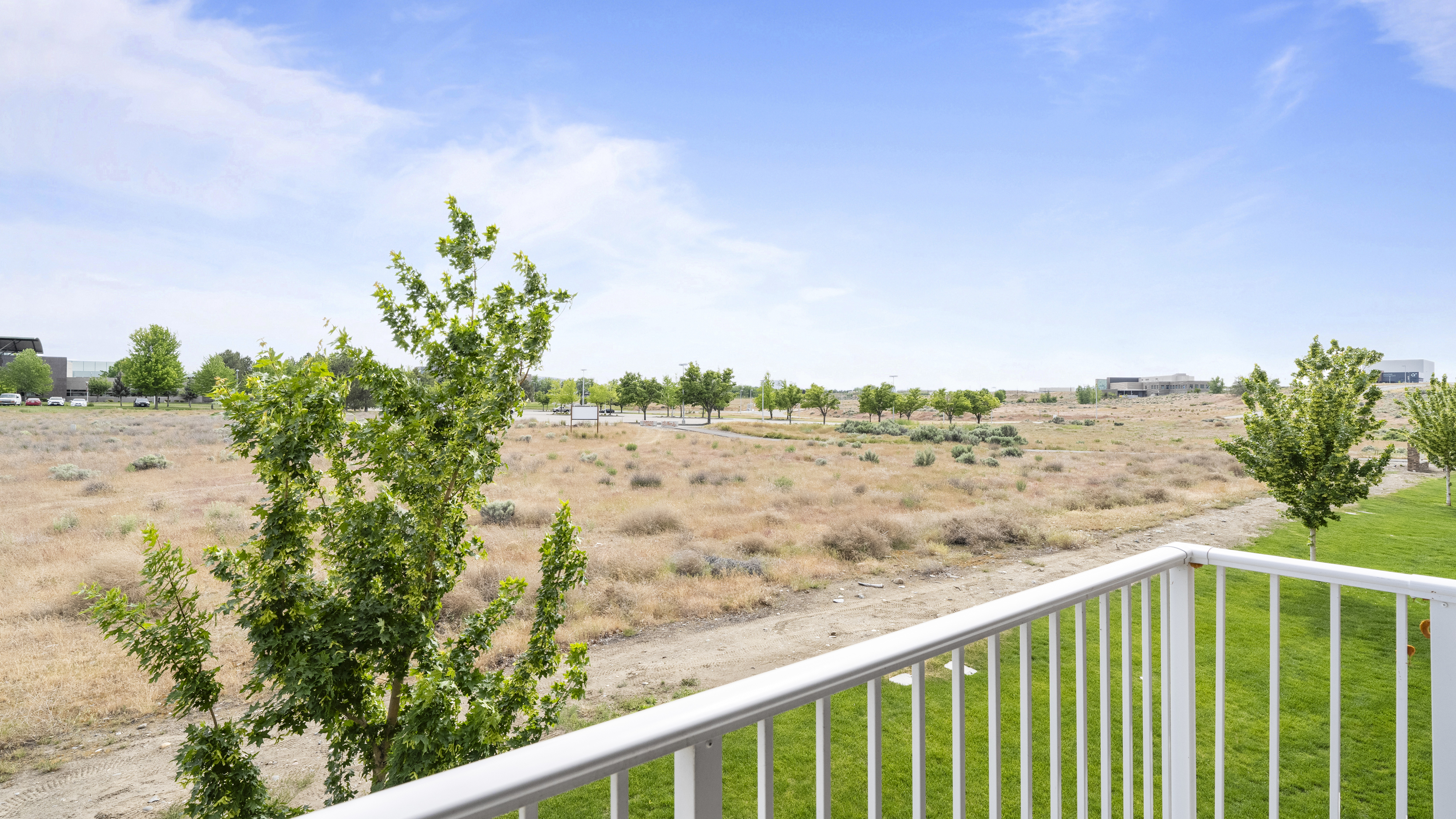 Bright and serene landscape view from a balcony overlooking a lush green lawn and expansive open fields under a clear blue sky.