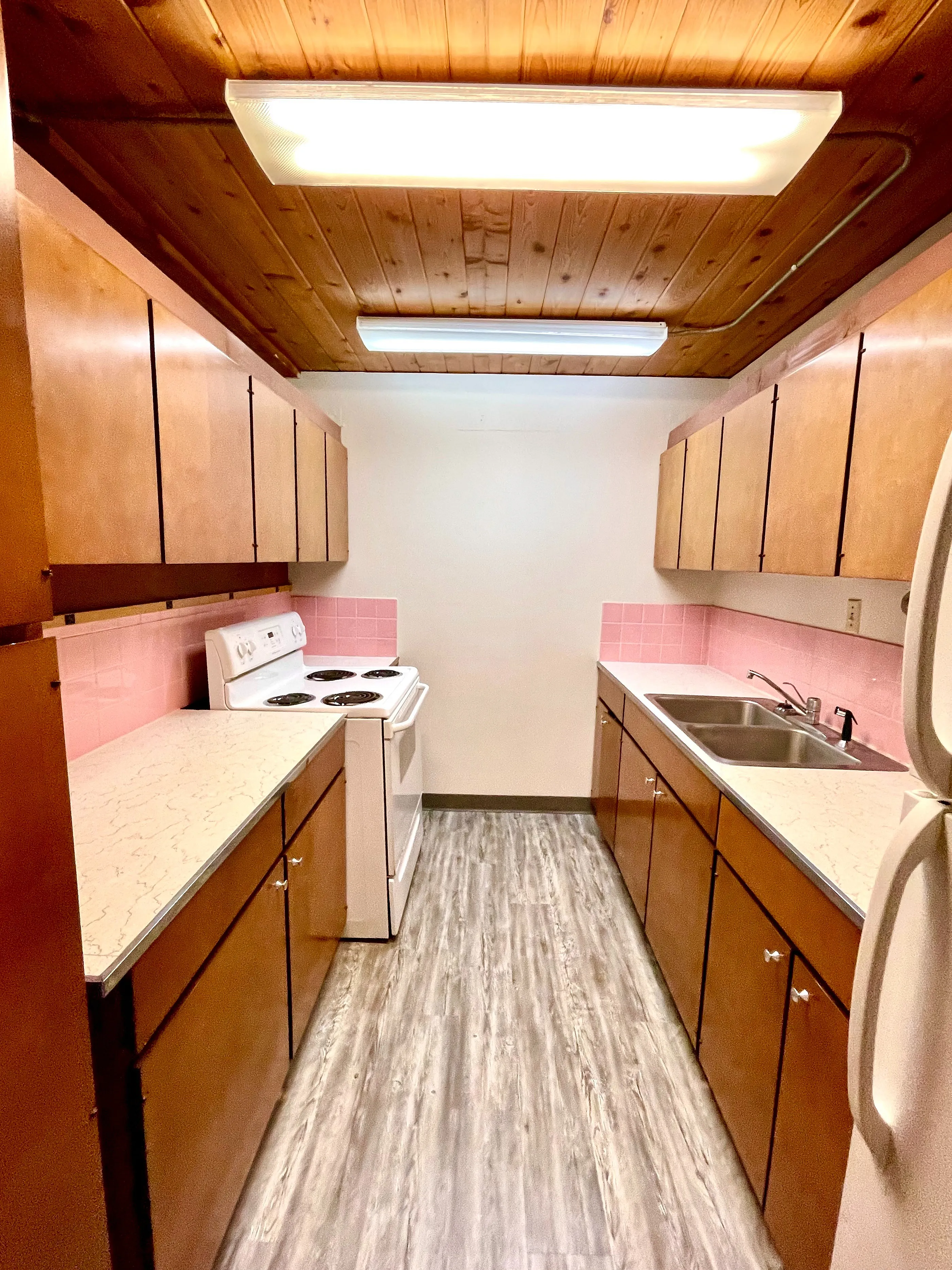 Charming galley kitchen featuring warm wood cabinetry, pink tile accents, and modern appliances under a stylish wood-paneled ceiling.