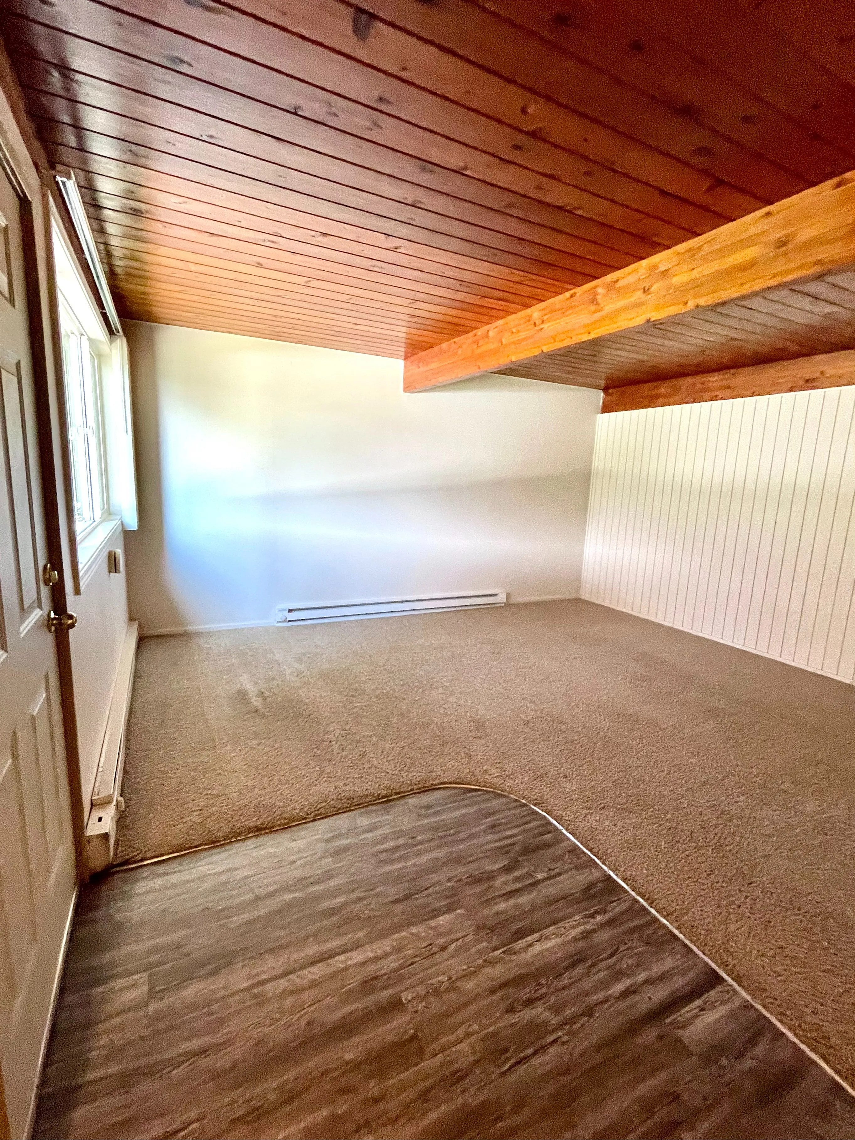 Inviting room with rich wood panel ceiling and cozy carpeted floor, featuring large window for natural light.