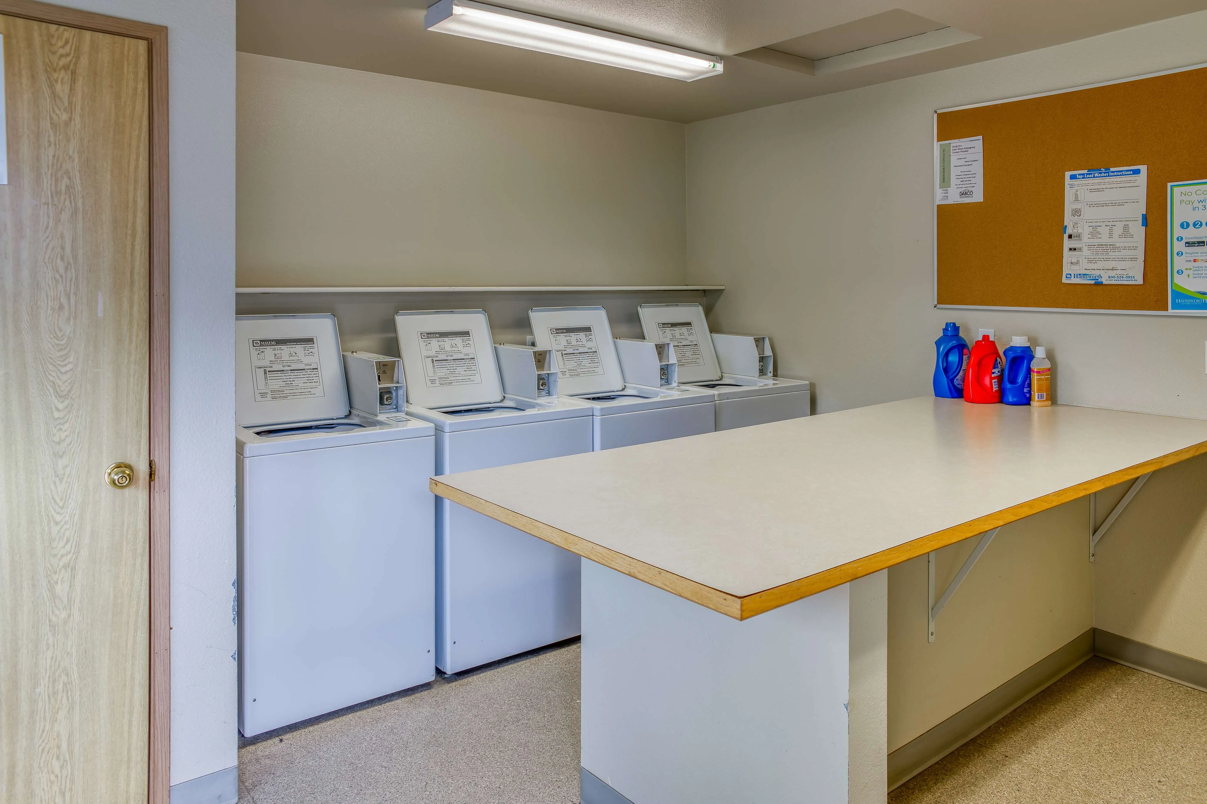 Modern laundry room with multiple washing machines and a spacious folding table, enhanced by bright natural lighting and organized detergent storage.