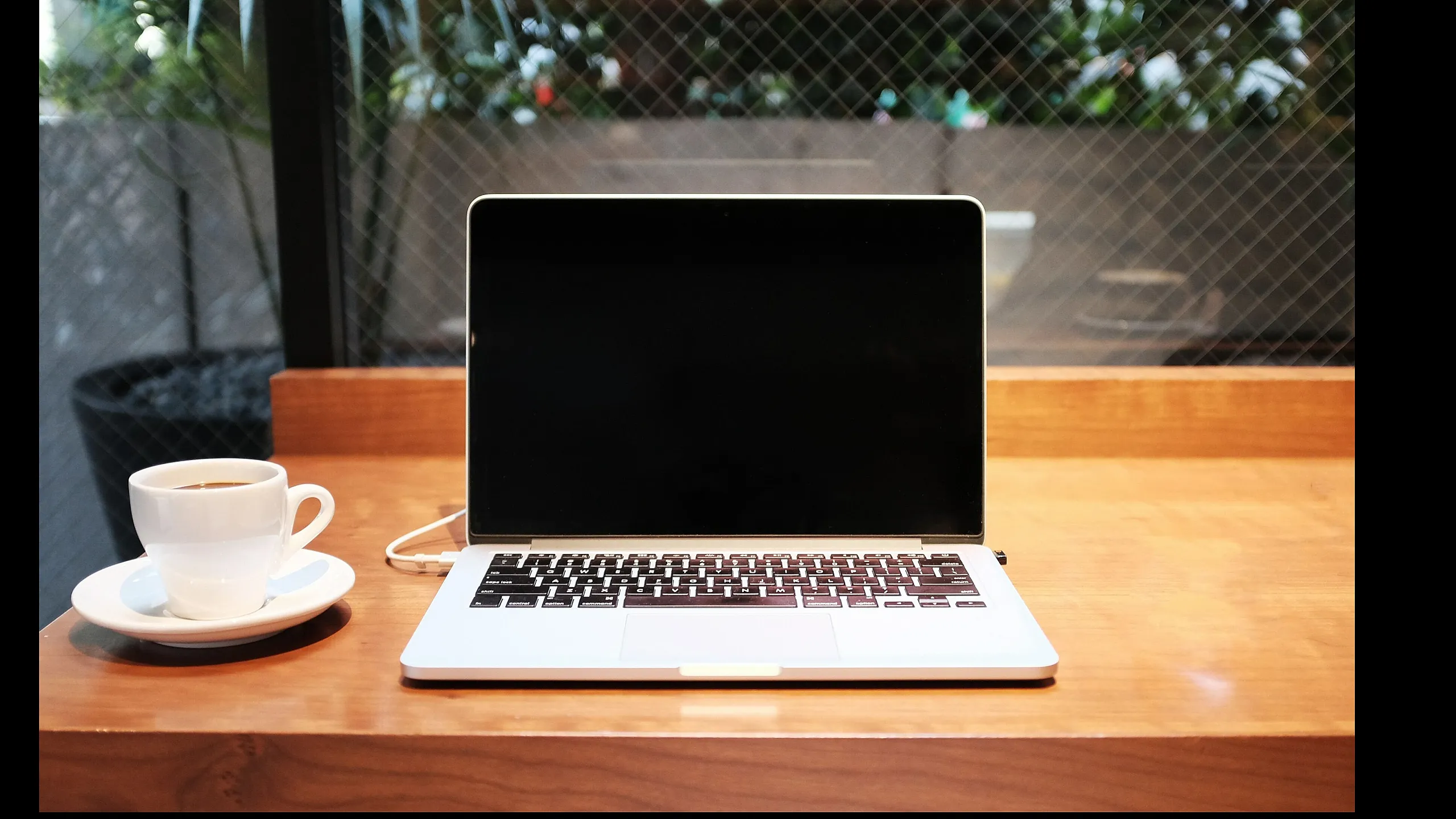 Cozy workspace with an elegant laptop and steaming cup of coffee on a wooden table by a large window, offering a serene view.