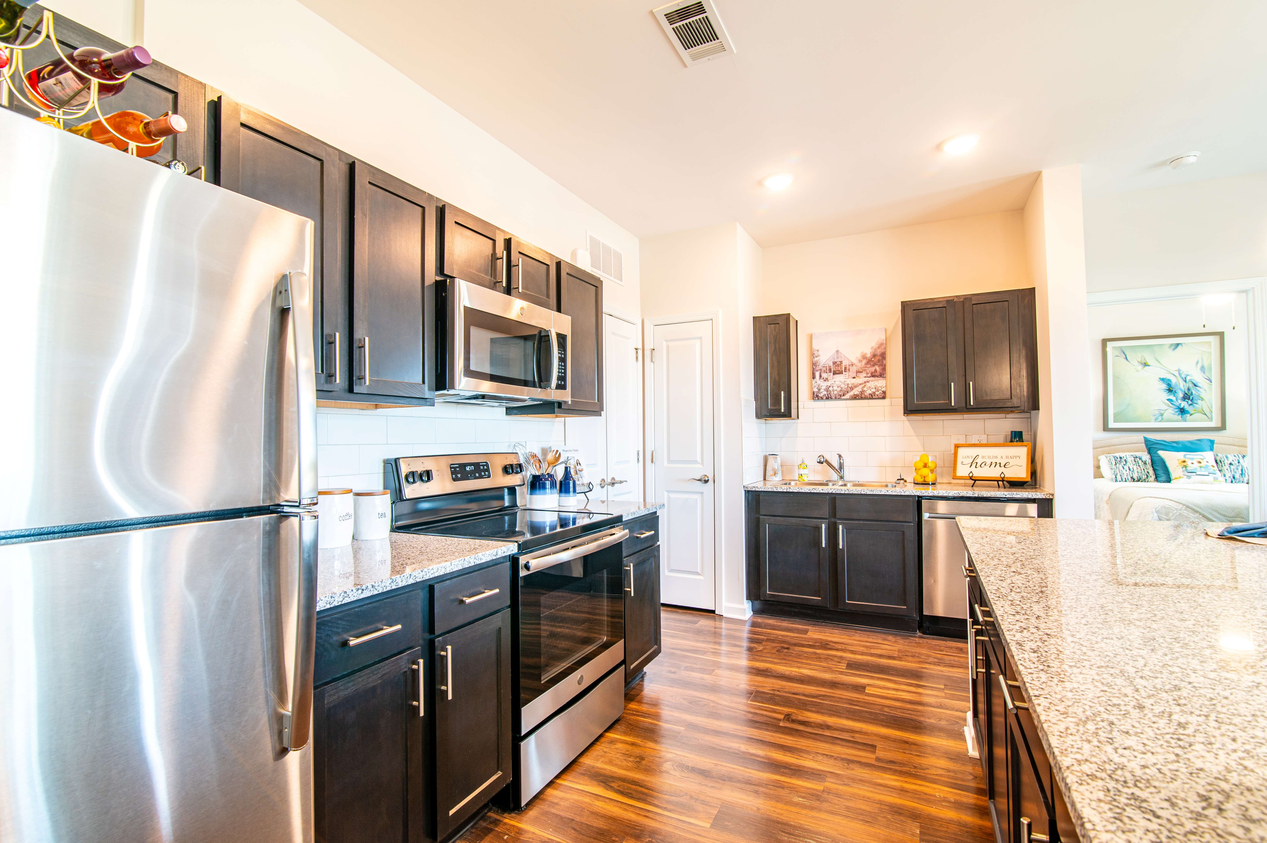 Modern kitchen with sleek dark cabinets, stainless steel appliances, wooden flooring, and a cozy open layout leading to a sunlit bedroom.