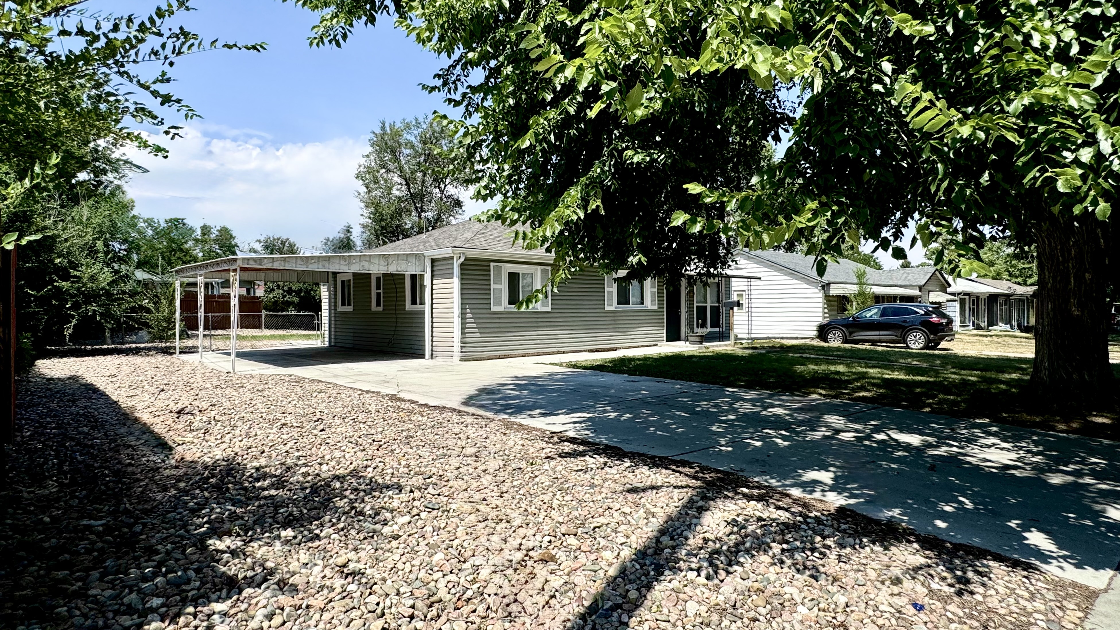 Charming single-story home with a covered carport and spacious gravel driveway, shaded by lush greenery.