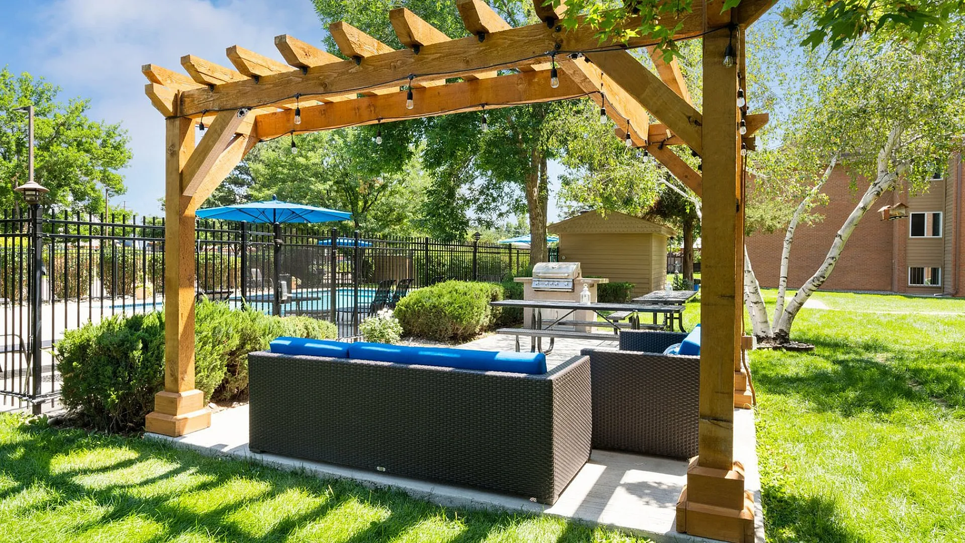 Charming outdoor patio with pergola, featuring cozy seating and a barbecue, set amidst lush greenery and a sparkling pool in the background.