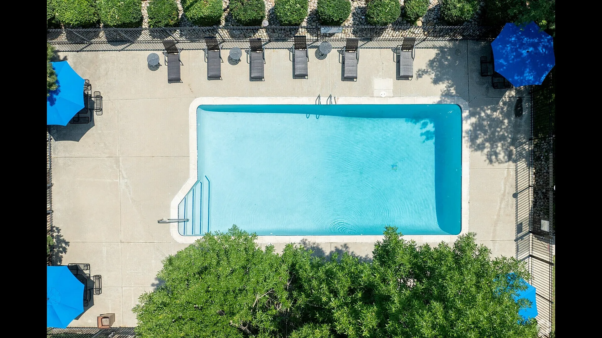 Inviting aerial view of a pristine swimming pool surrounded by lush greenery and elegant lounge chairs with blue umbrellas, perfect for luxurious relaxation.