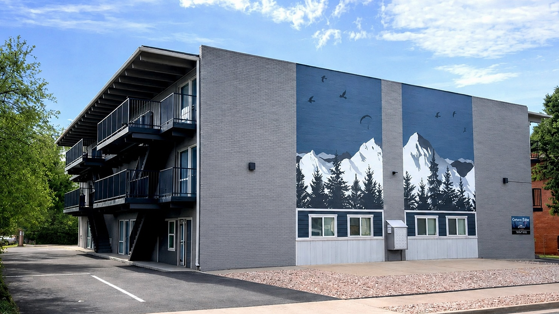 Modern apartment building with striking mural of mountains and birds, featuring stylish balconies and surrounded by lush greenery.