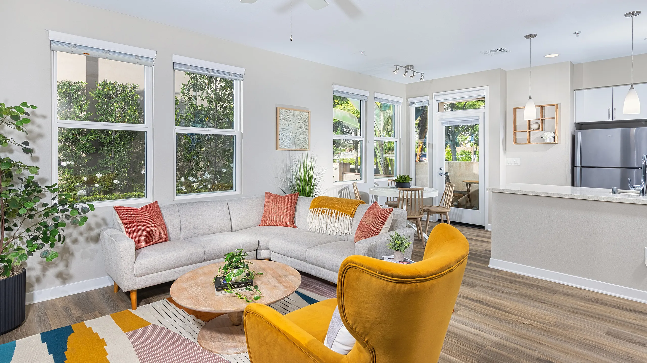 Bright and inviting living area with cozy gray sectional, stylish yellow armchair, and natural light streaming through large windows, leading to a modern open kitchen.