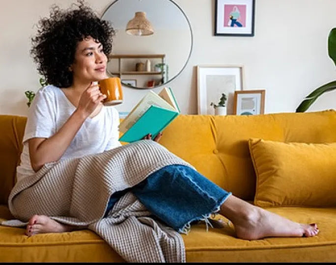 Cozy living room scene with a cheerful person relaxing on a trendy yellow sofa, enjoying a book and a warm drink, surrounded by modern decor and lush greenery.