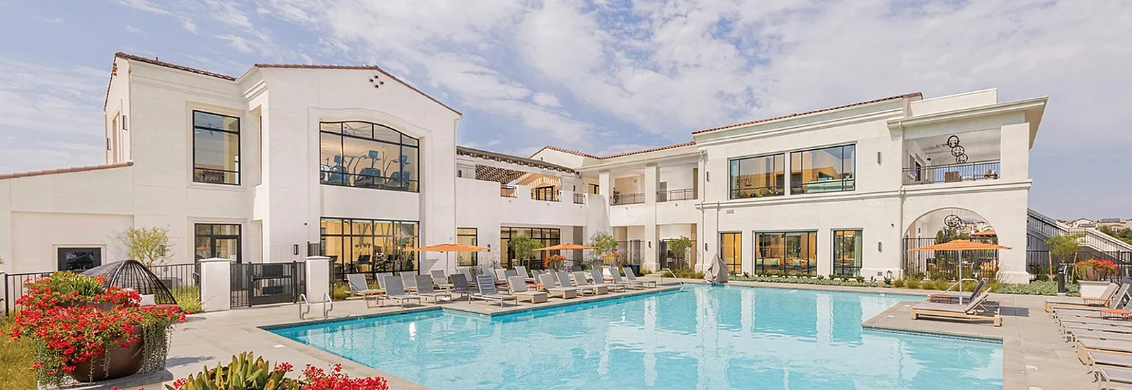 Luxurious apartment community with a pristine swimming pool surrounded by stylish lounge chairs and vibrant flowers, set against a clear blue sky.
