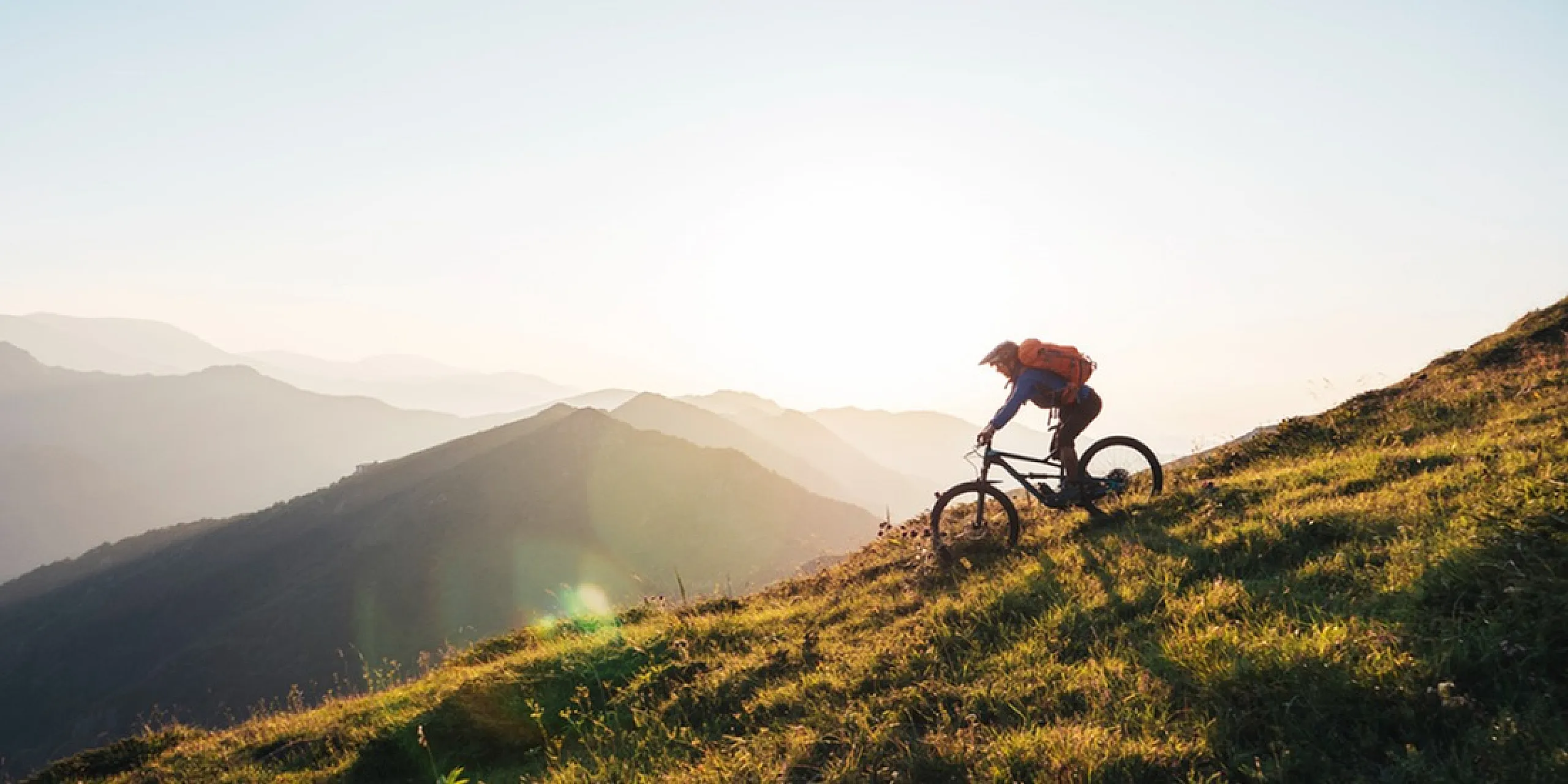Adventurous mountain biker descending a sunlit grassy hill, surrounded by breathtaking scenic mountain views.