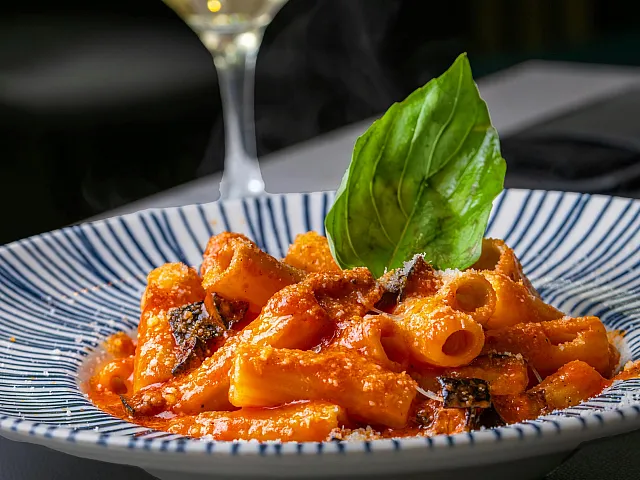 Elegant dining setup features a steaming plate of rigatoni with rich tomato sauce, garnished with a fresh basil leaf, paired perfectly with a glass of white wine.