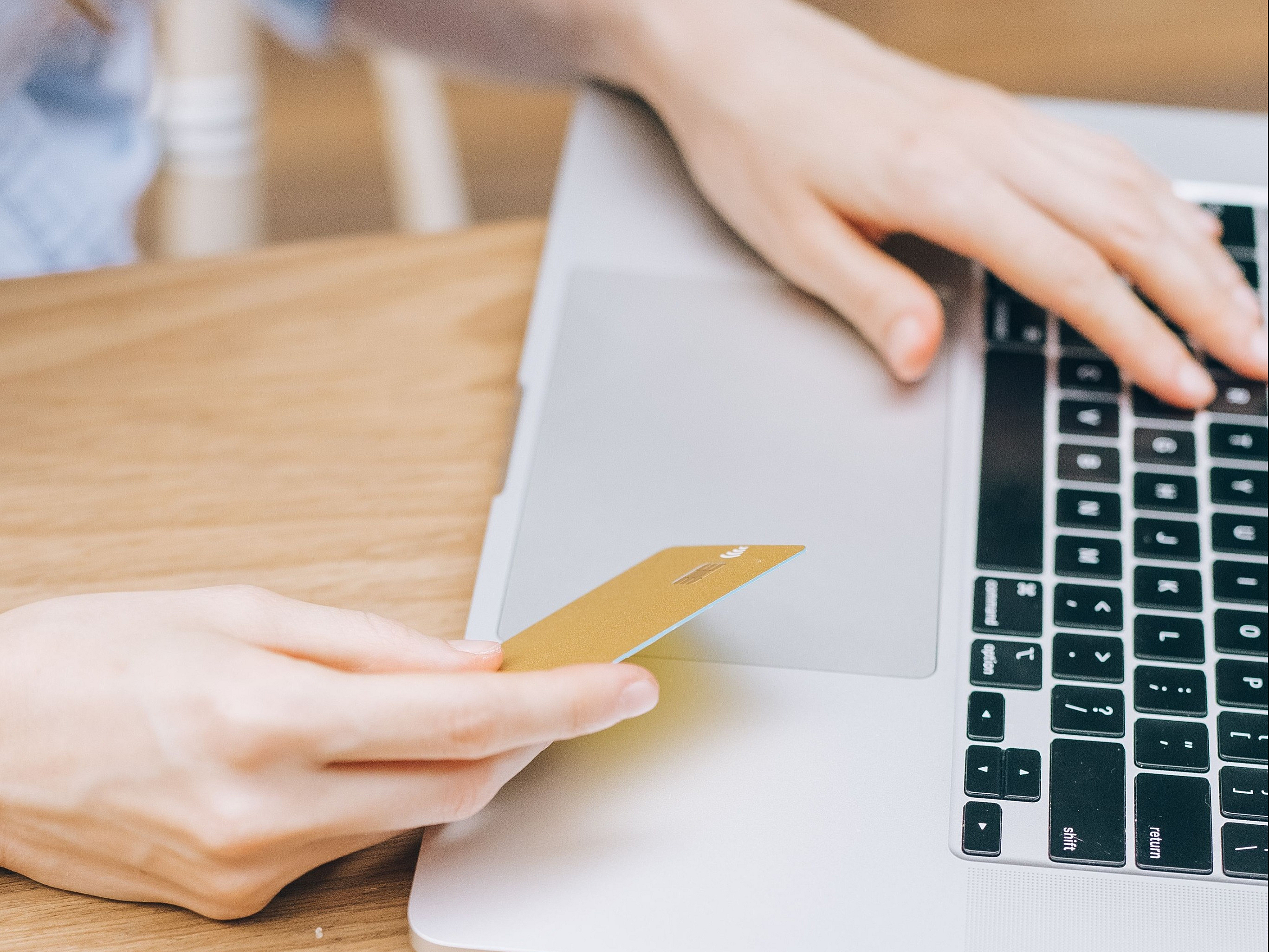 Close-up of a person holding a gold credit card while using a laptop, with a stylish pair of eyeglasses and a cup of coffee nearby on a wooden table, signifying modern online shopping convenience.