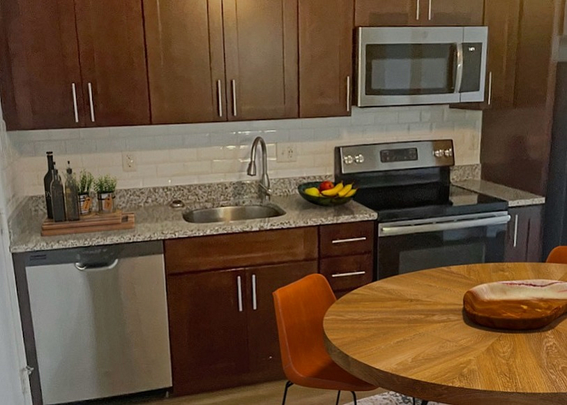 Contemporary kitchen and living area featuring sleek dark wood cabinetry, stainless steel appliances, a round wooden dining table with modern chairs, and a cozy, stylishly decorated living room.