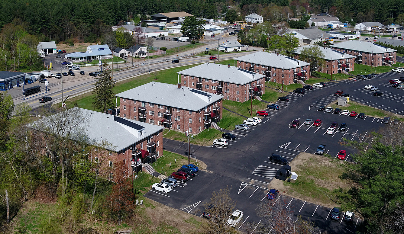 Charming aerial view of a serene apartment complex with multiple brick buildings, ample parking, and lush greenery, ideal for convenient and peaceful living.