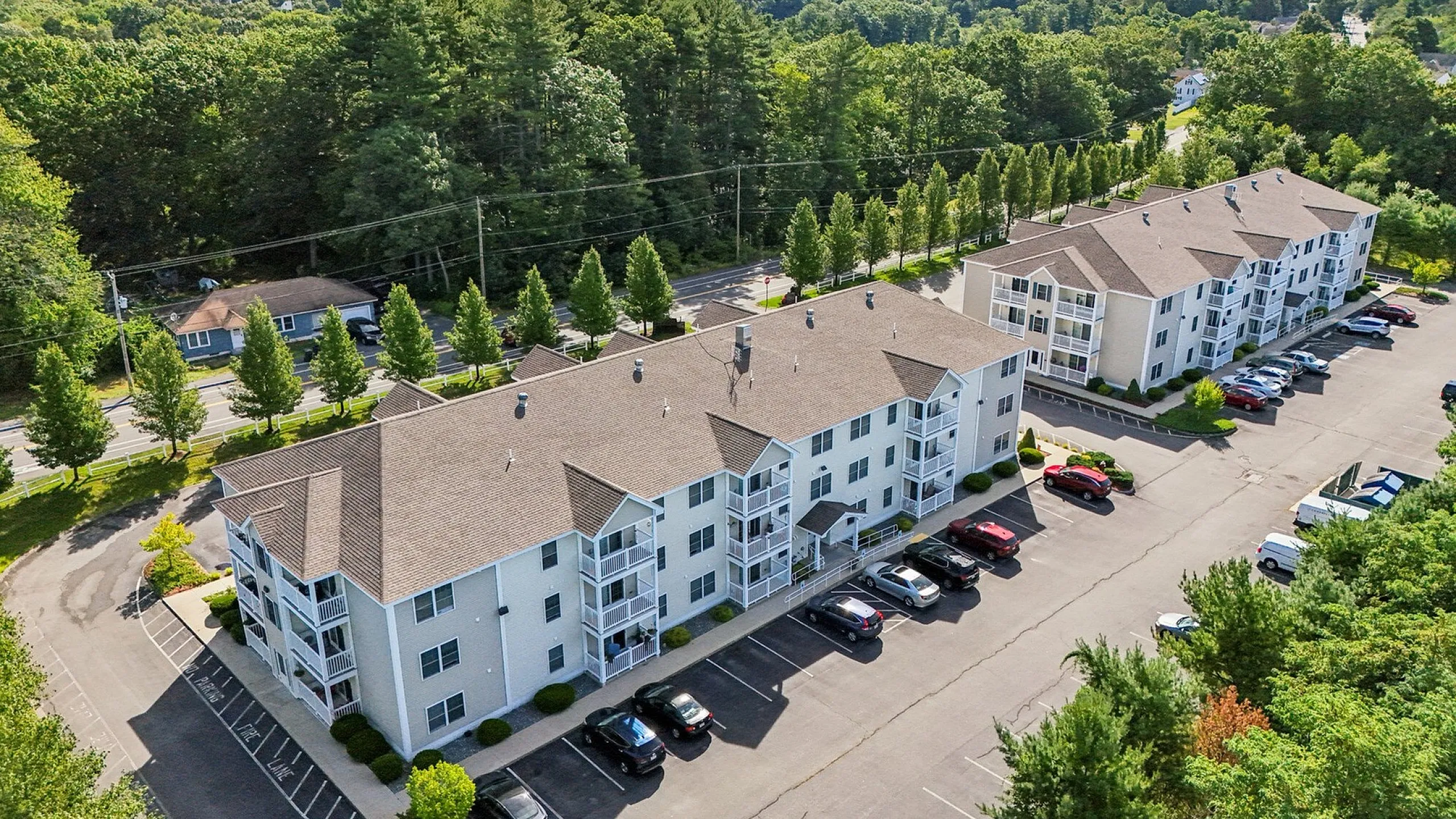 Aerial view of charming apartment complex surrounded by lush greenery, featuring spacious parking and serene residential setting.
