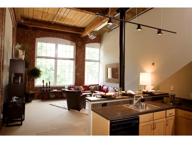 Apartment interior view with corner of kitchen and high-top counter visible in foreground. Living room with oversized windows, exposed brick and 16\' ceiling with exposed beams in background.