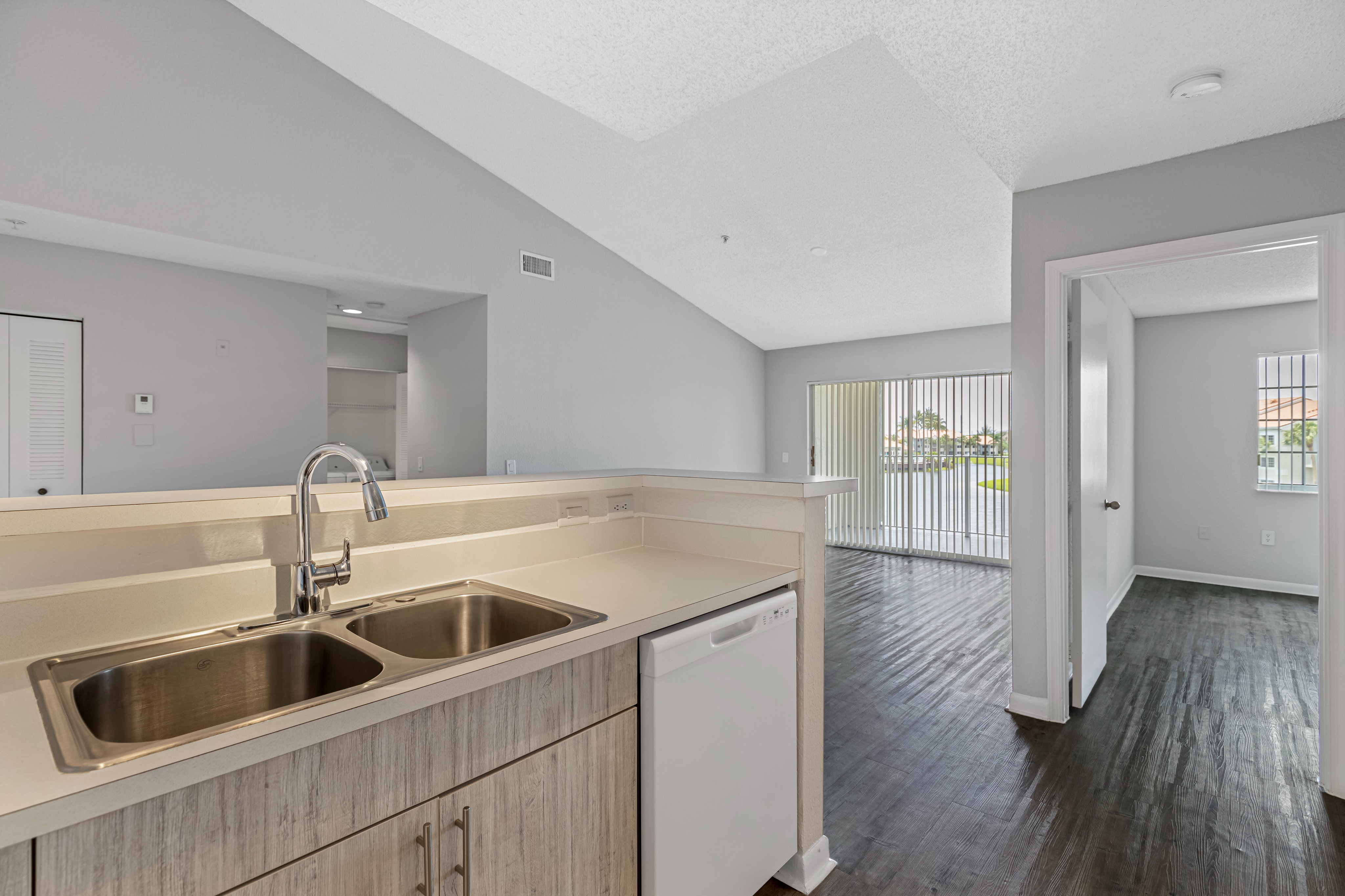 Apartment kitchen with white refrigerator and dishwasher connected to unfurnished living room with wood floors, vaulted ceiling, tracking lighting, sliding glass balcony doors, and raised bar peninsula countertop