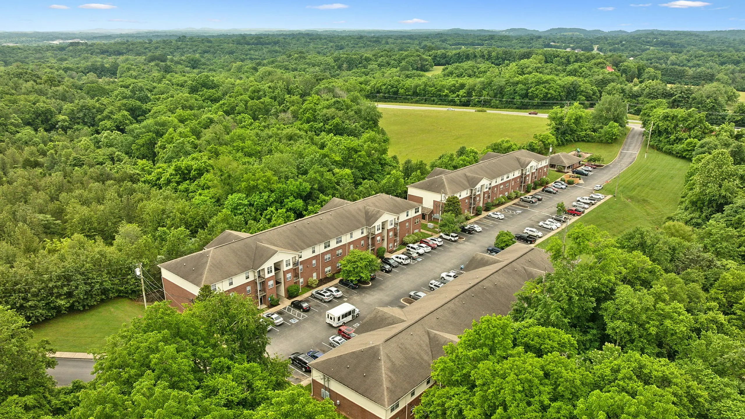 Aerial view of a charming apartment complex surrounded by lush green forest, showcasing ample parking and serene natural beauty.