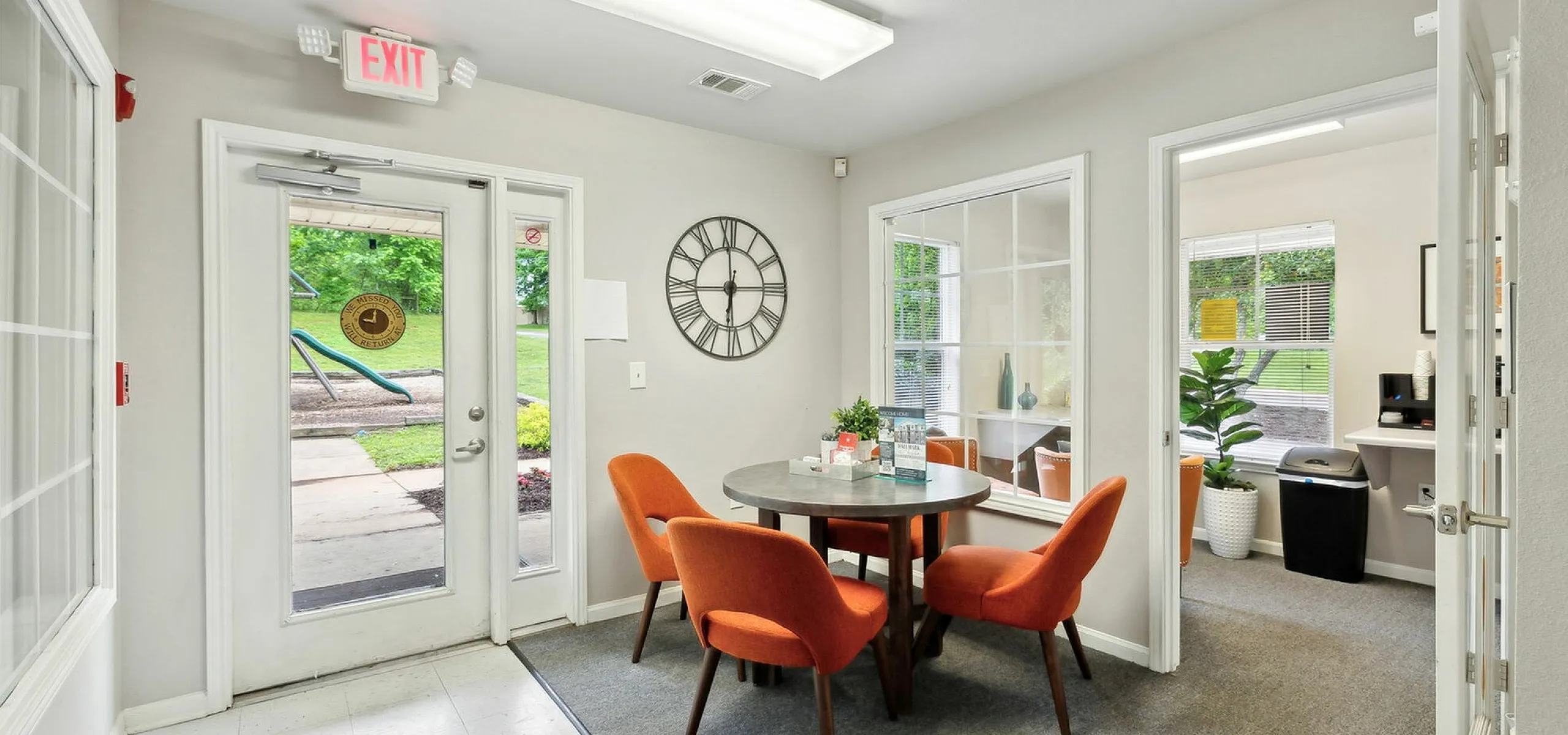 Charming office space featuring vibrant orange chairs around a sleek round table, with large windows offering a view of lush greenery.