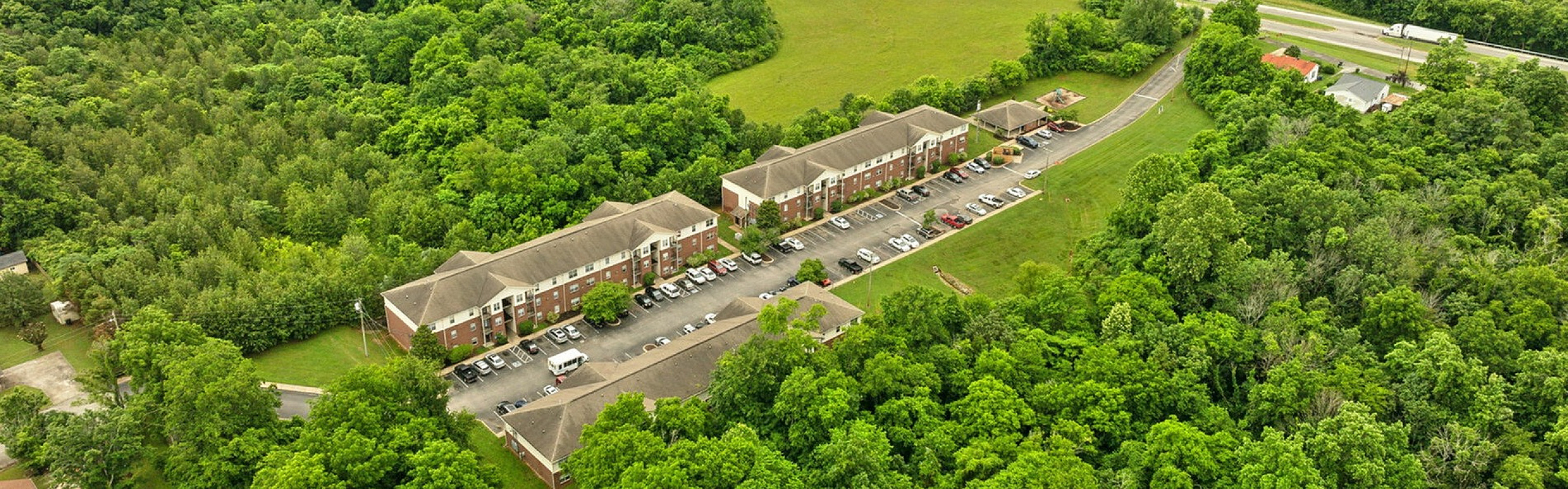 Aerial view of charming apartment complex nestled in lush green landscape, offering serene living with convenient highway access.