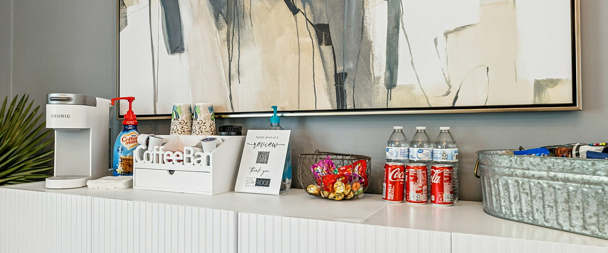 Inviting coffee bar setup with modern Keurig machine, decorative cups, and an array of snacks and beverages, set against a chic abstract art backdrop.